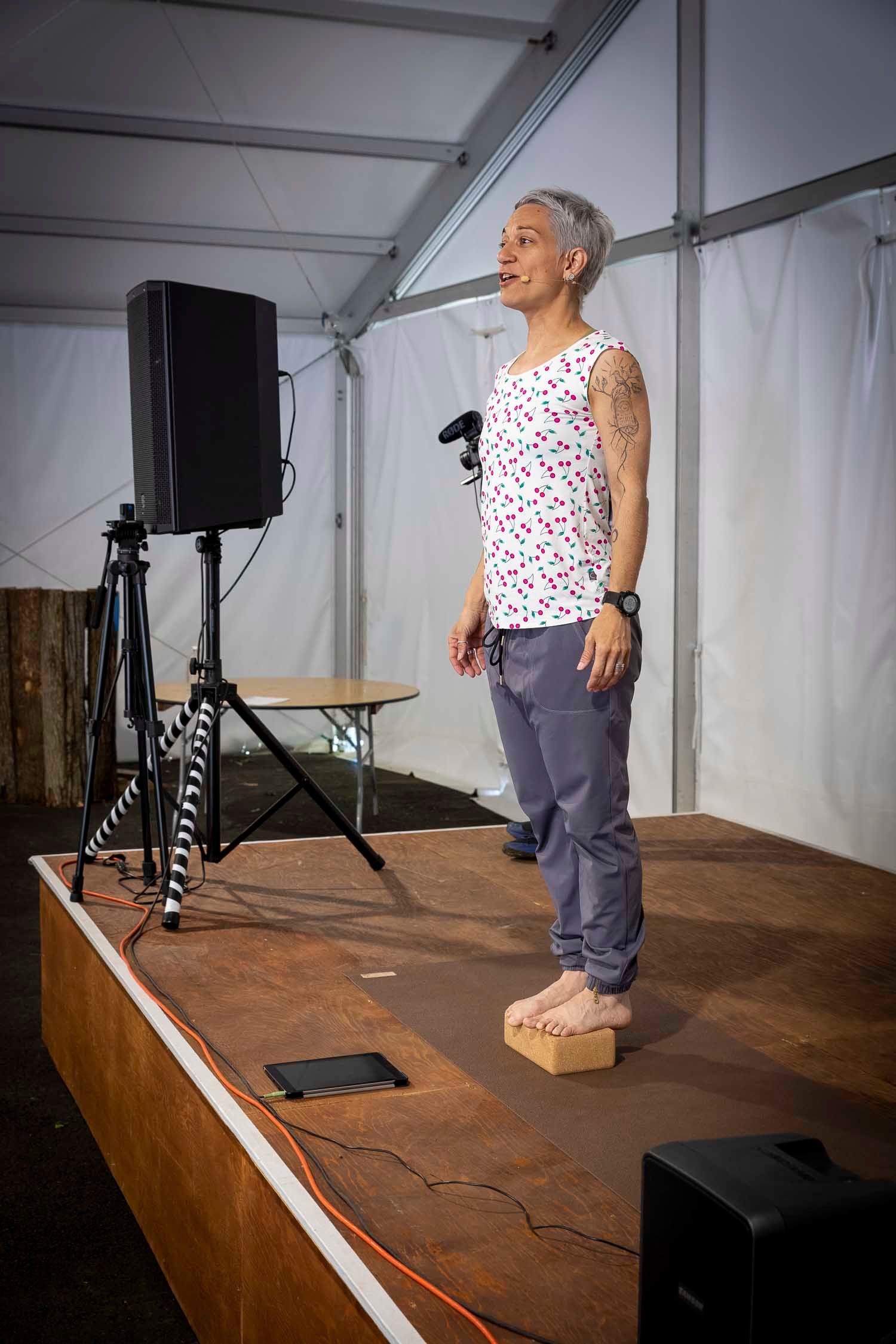 Woman with tattoos stands on stage in floral tank top and gray pants, speaking at an outdoor event under a tent.