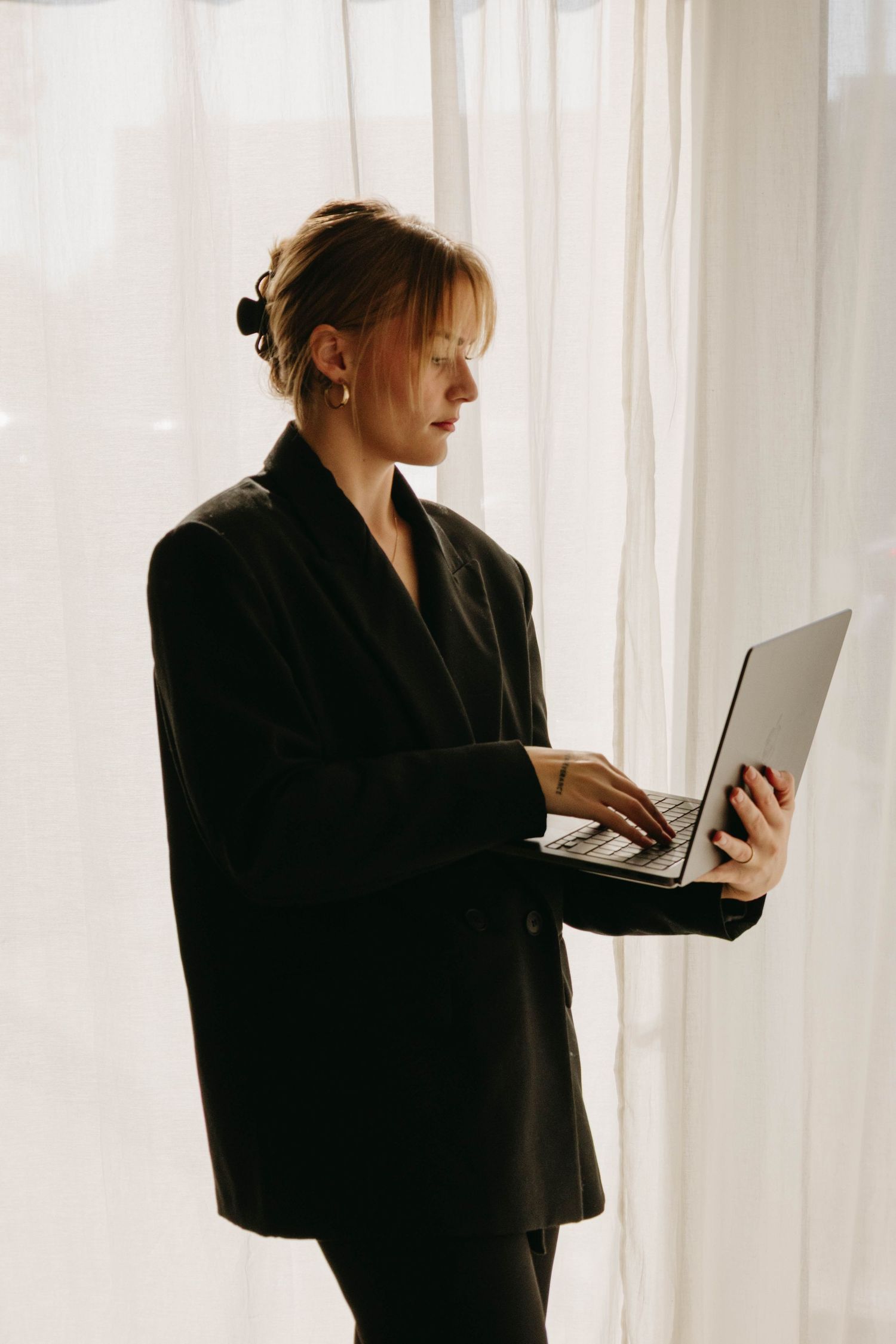Professional woman in black blazer standing by window using a laptop computer indoors.