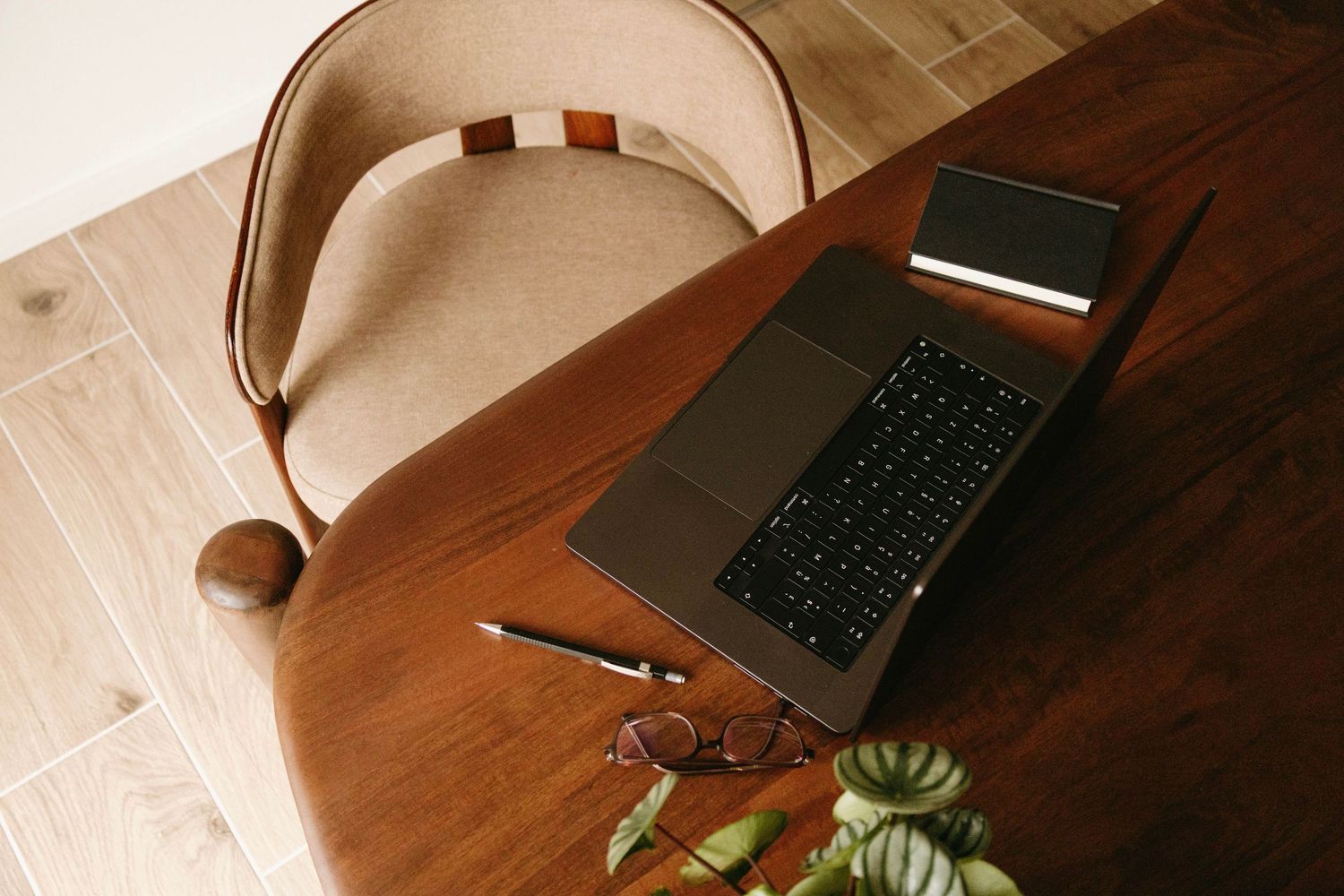 Overhead view of open laptop, glasses, pen, and plant on a wooden desk with chair.