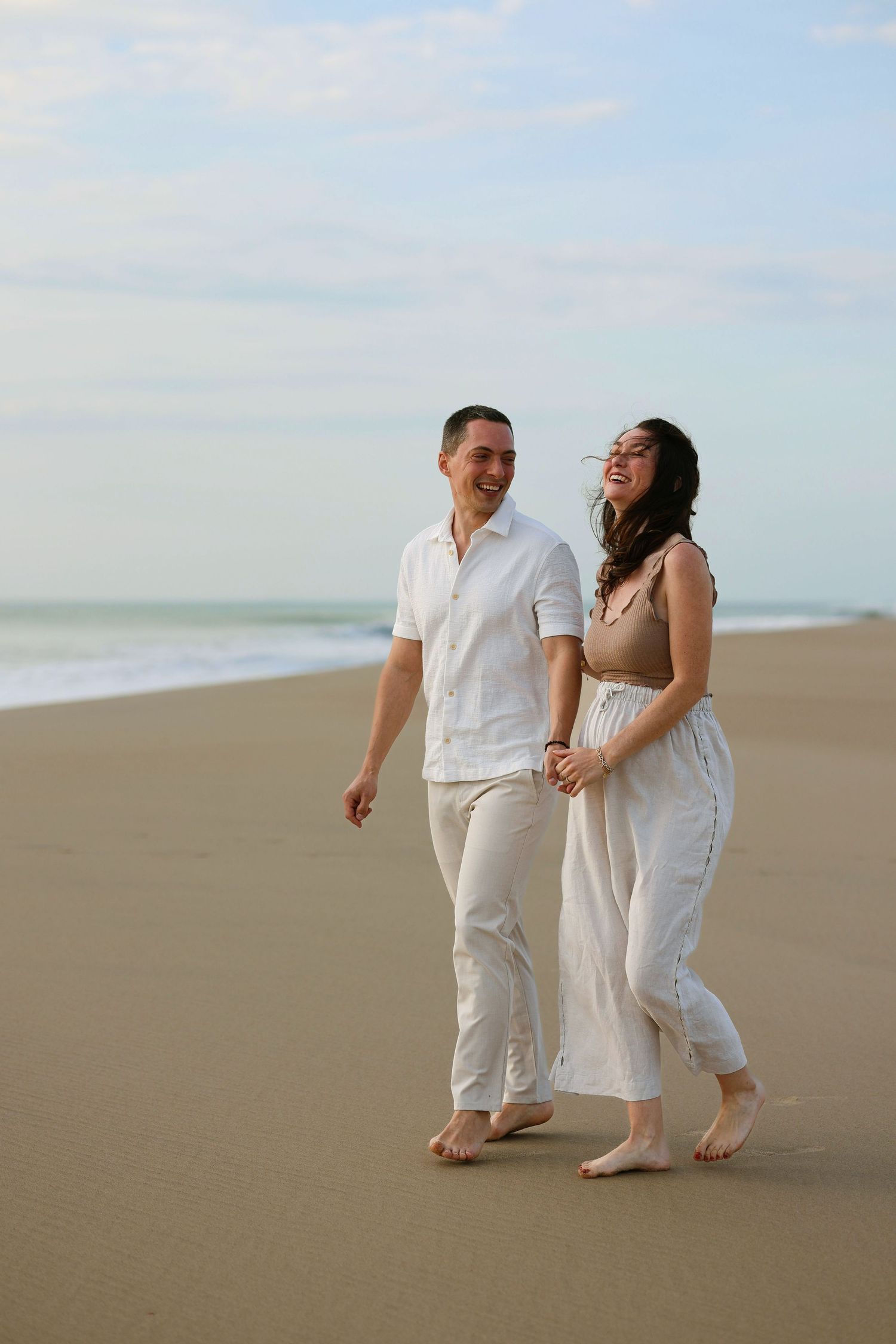 Happy couple holding hands and walking barefoot on a sandy beach in white outfits.