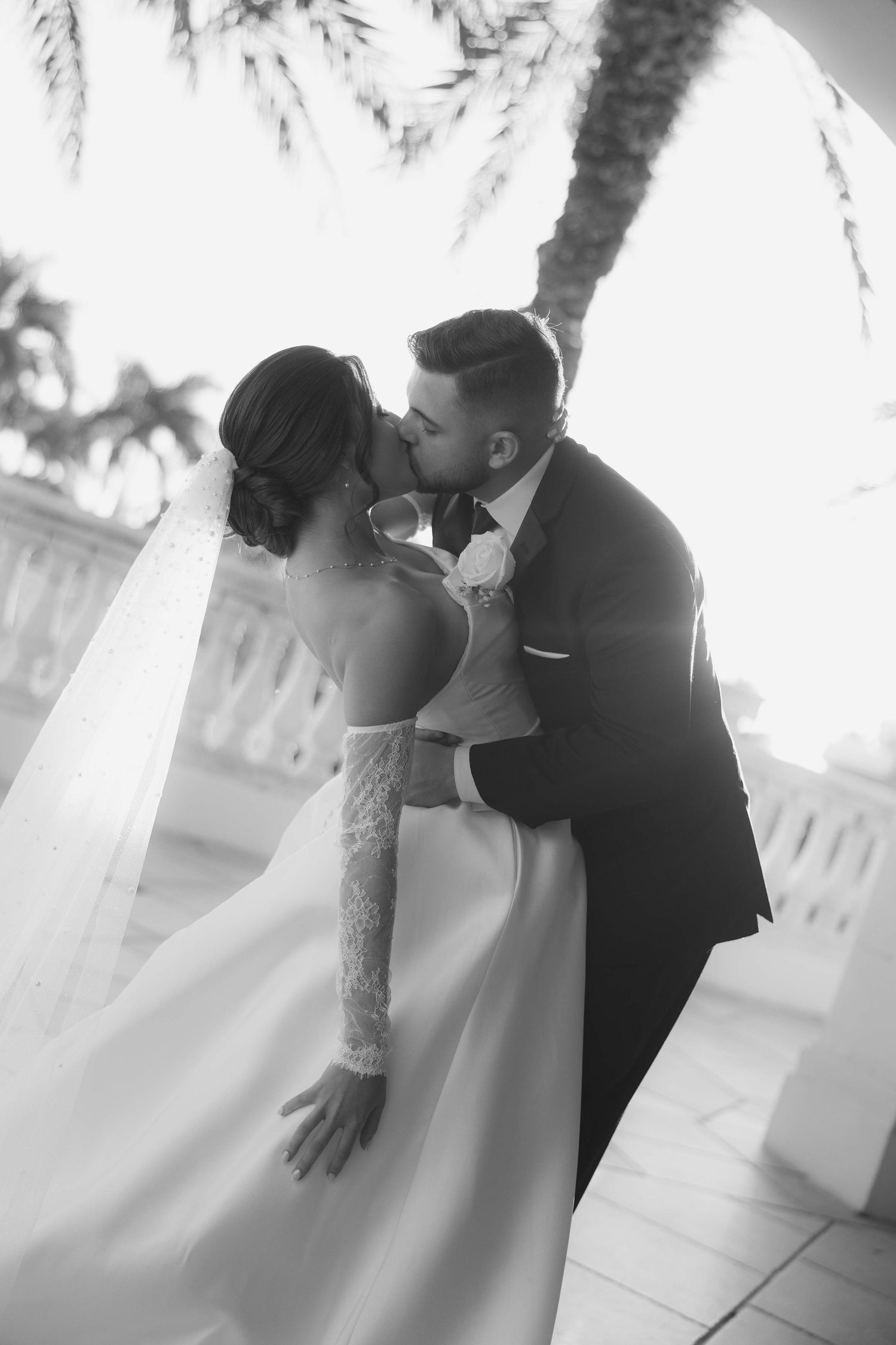 Romantic black and white wedding photo of newlyweds sharing intimate kiss under palm trees.