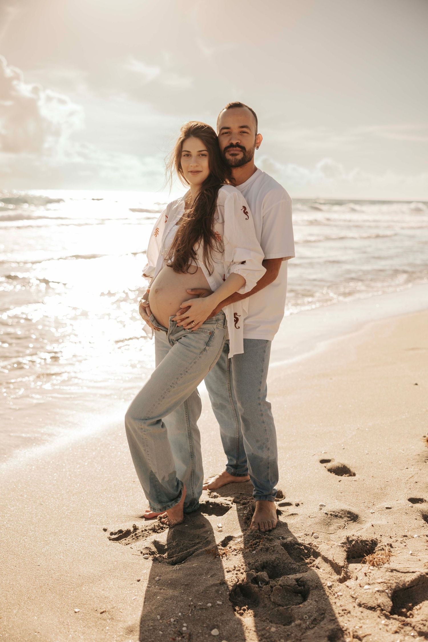 A couple stands together on a sandy beach during sunset with waves crashing behind them.