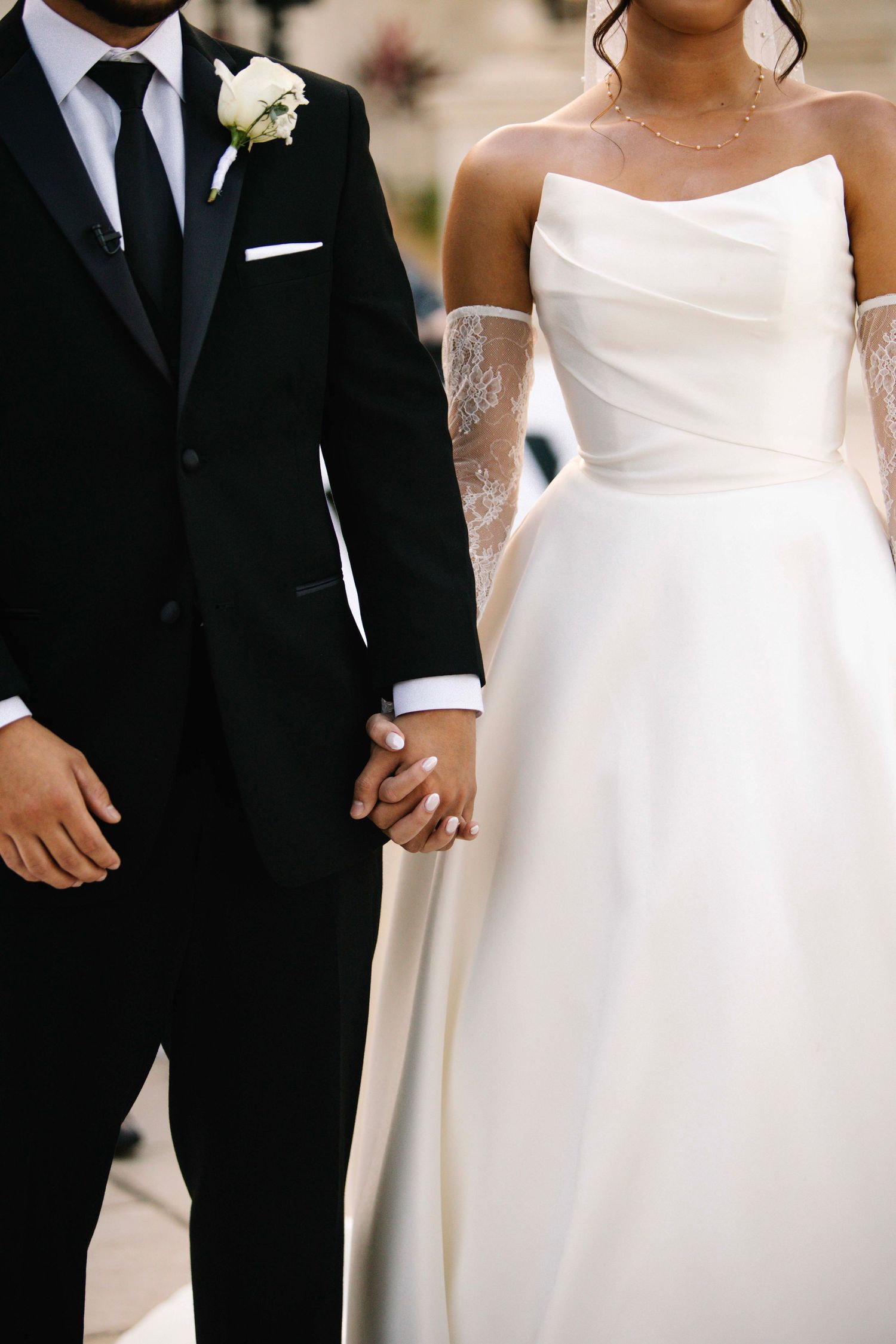 Close-up of a bride and groom holding hands, showing her white gown and his black tuxedo.