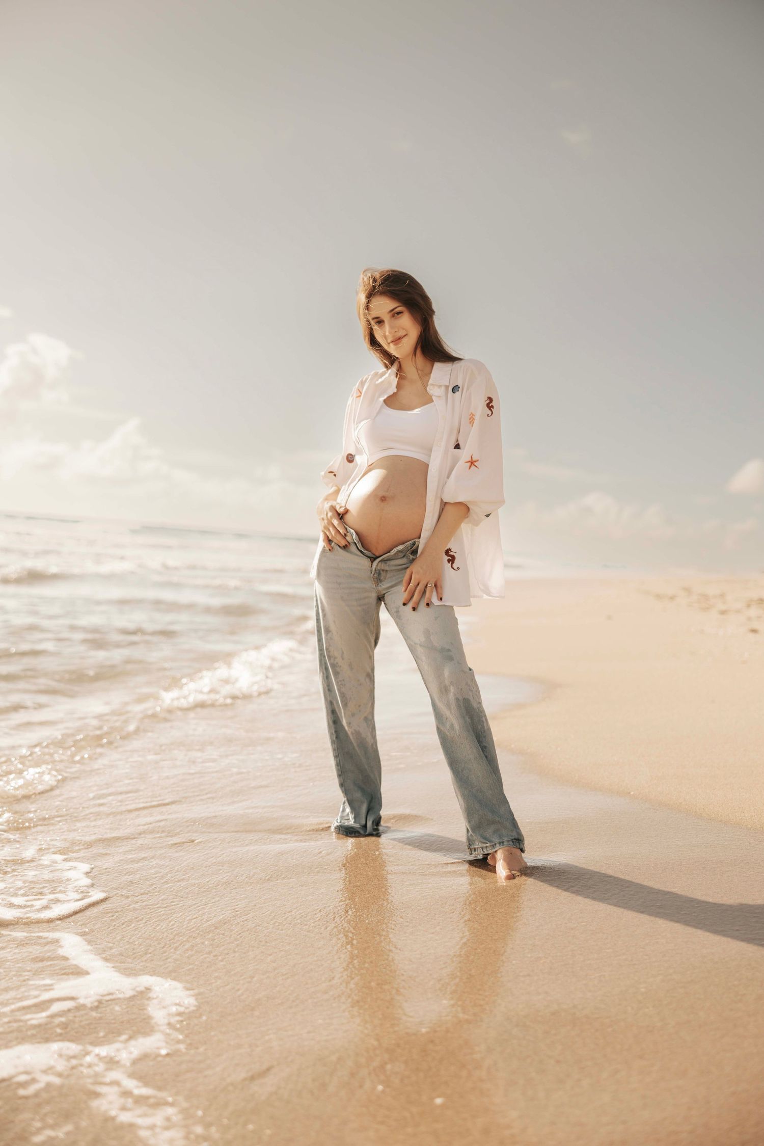 An expectant mother stands barefoot on a beach at sunset wearing casual denim.