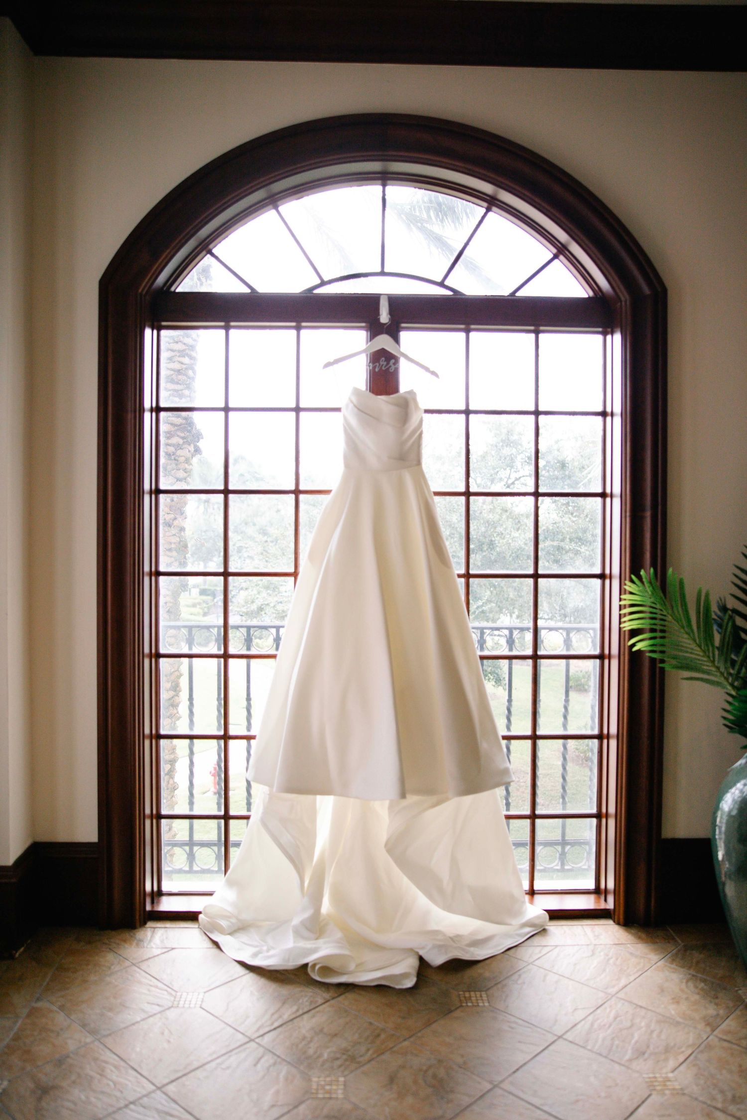 Elegant white wedding gown hanging in front of arched window with natural lighting.