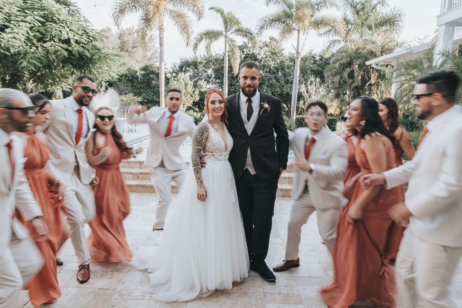 Wedding party poses outdoors with bridesmaids in coral dresses and groomsmen in light suits.