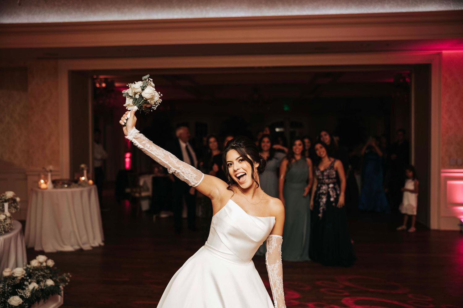 A joyful bride in a white gown celebrates on the dance floor with raised bouquet.