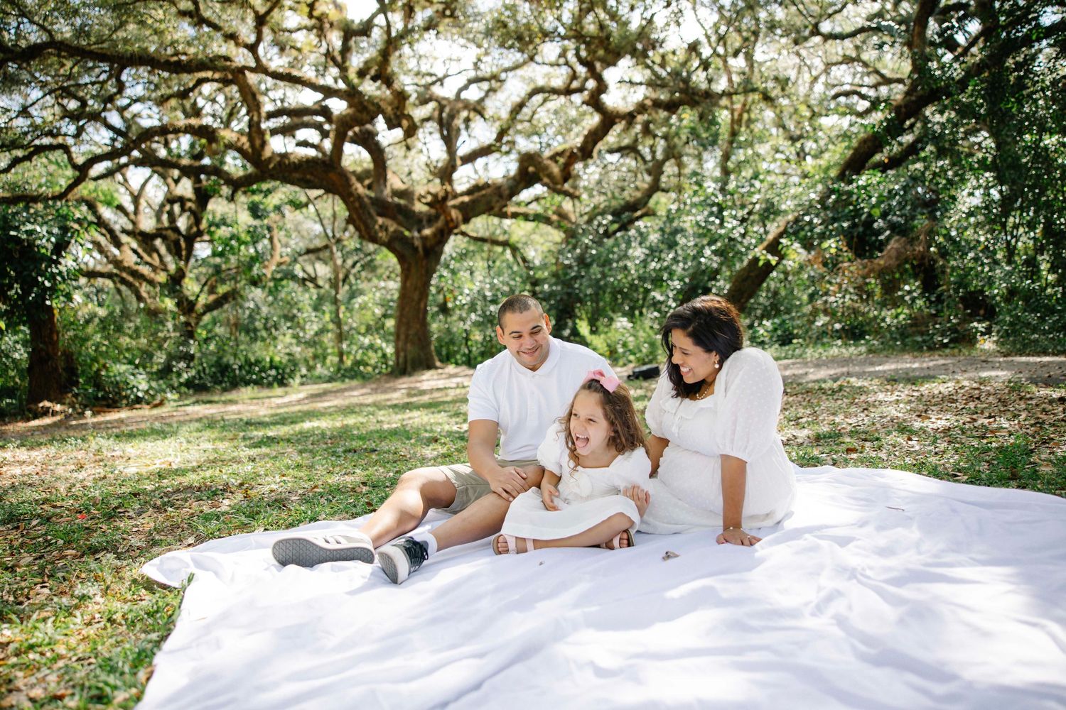 Family of three relaxing on white blanket under majestic oak tree.