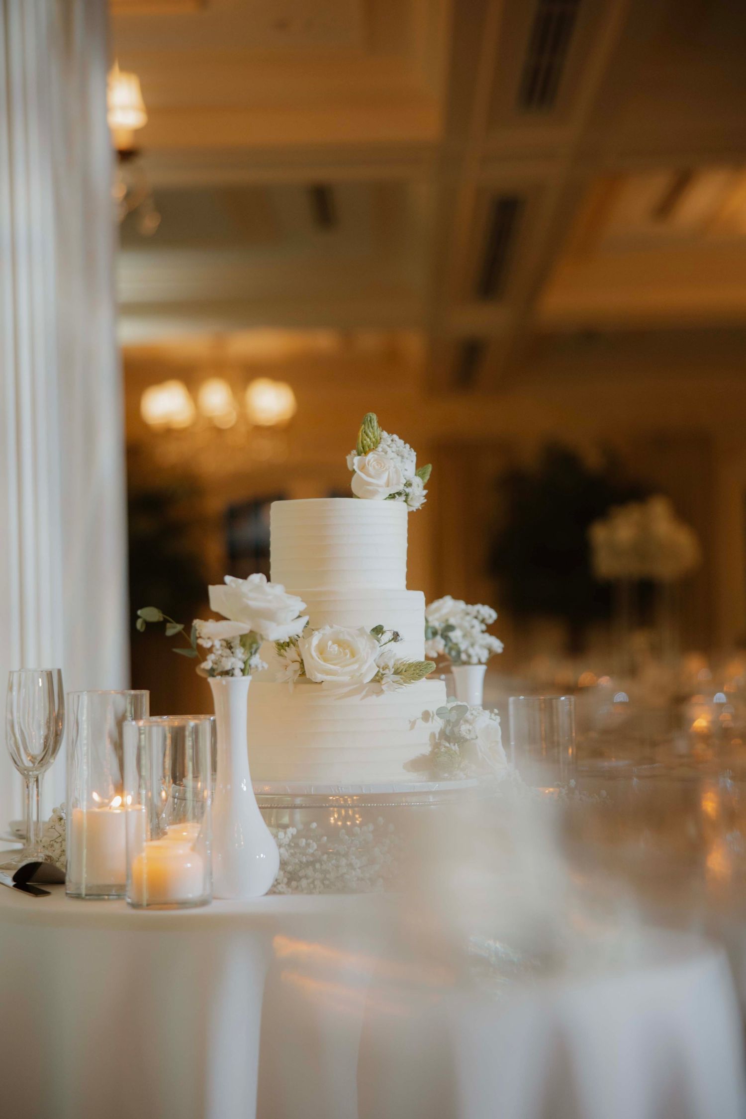 Elegant white tiered wedding cake with roses and candle decorations on reception table.