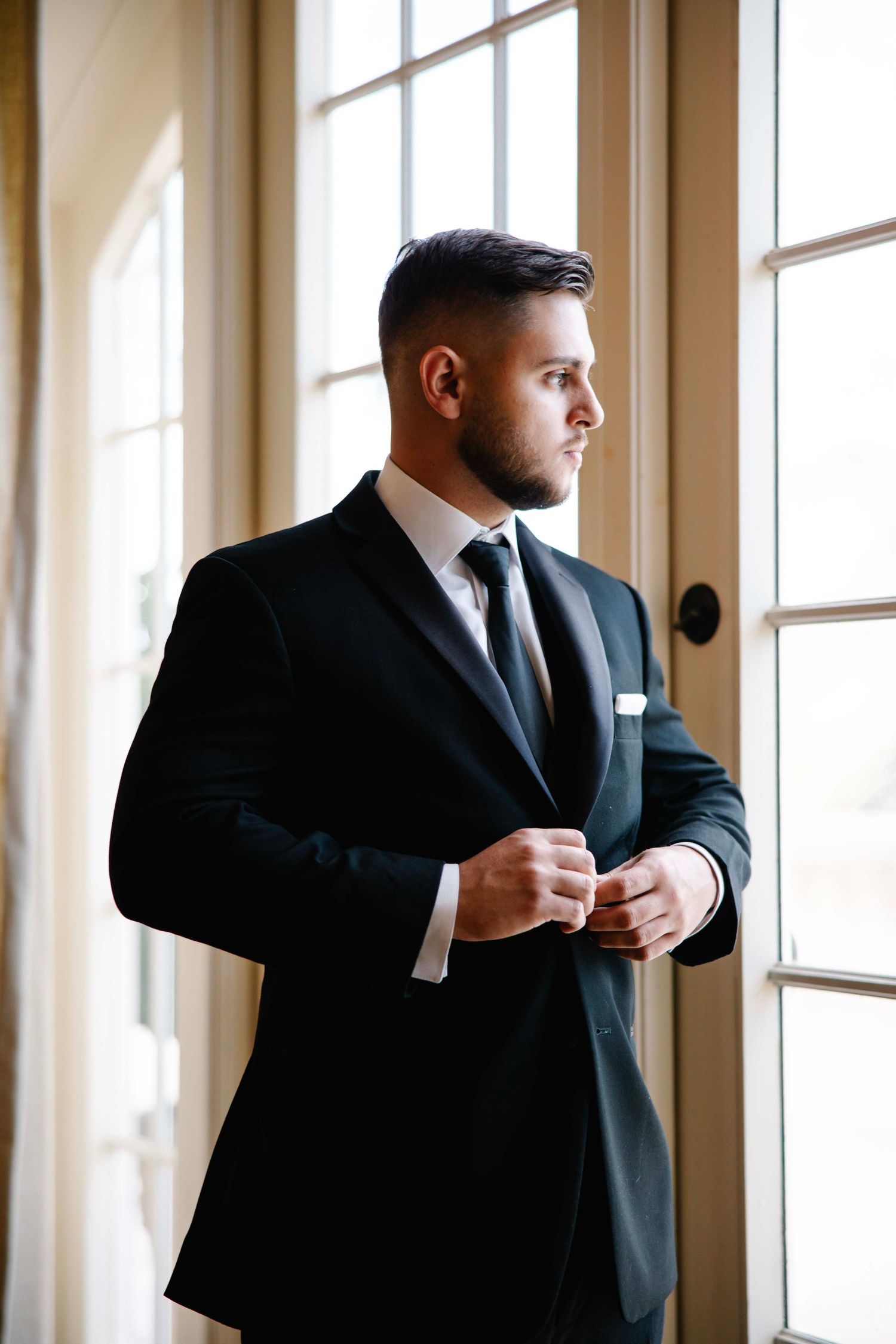 Groom in classic black suit adjusts cuff while standing by sunlit window.