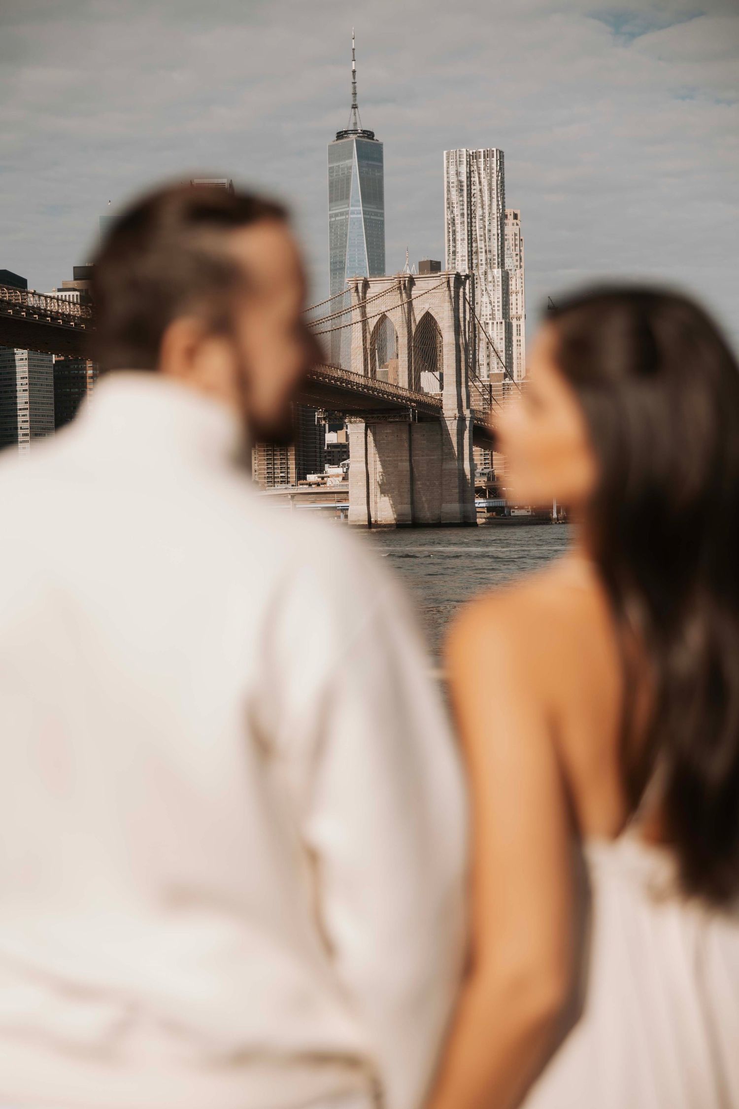 A couple gazes at the Brooklyn Bridge and One World Trade Center against a cloudy New York City skyline.
