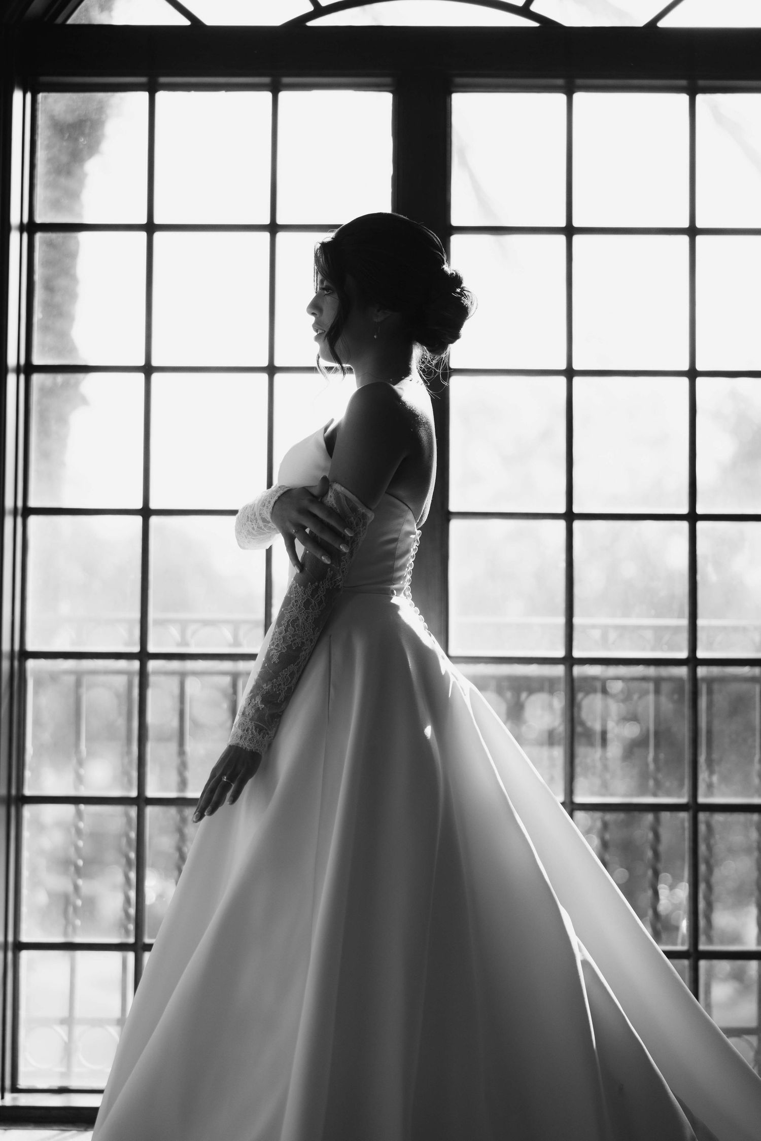 Silhouette of bride in ball gown wedding dress posed against large windows.