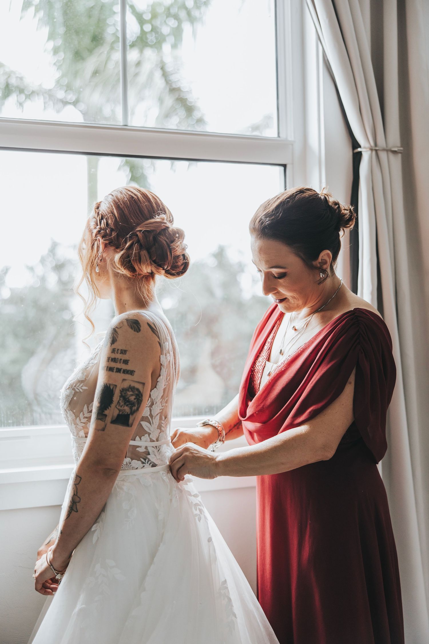 A bride in a white dress shows her tattoo while getting ready with help from someone in a red dress.