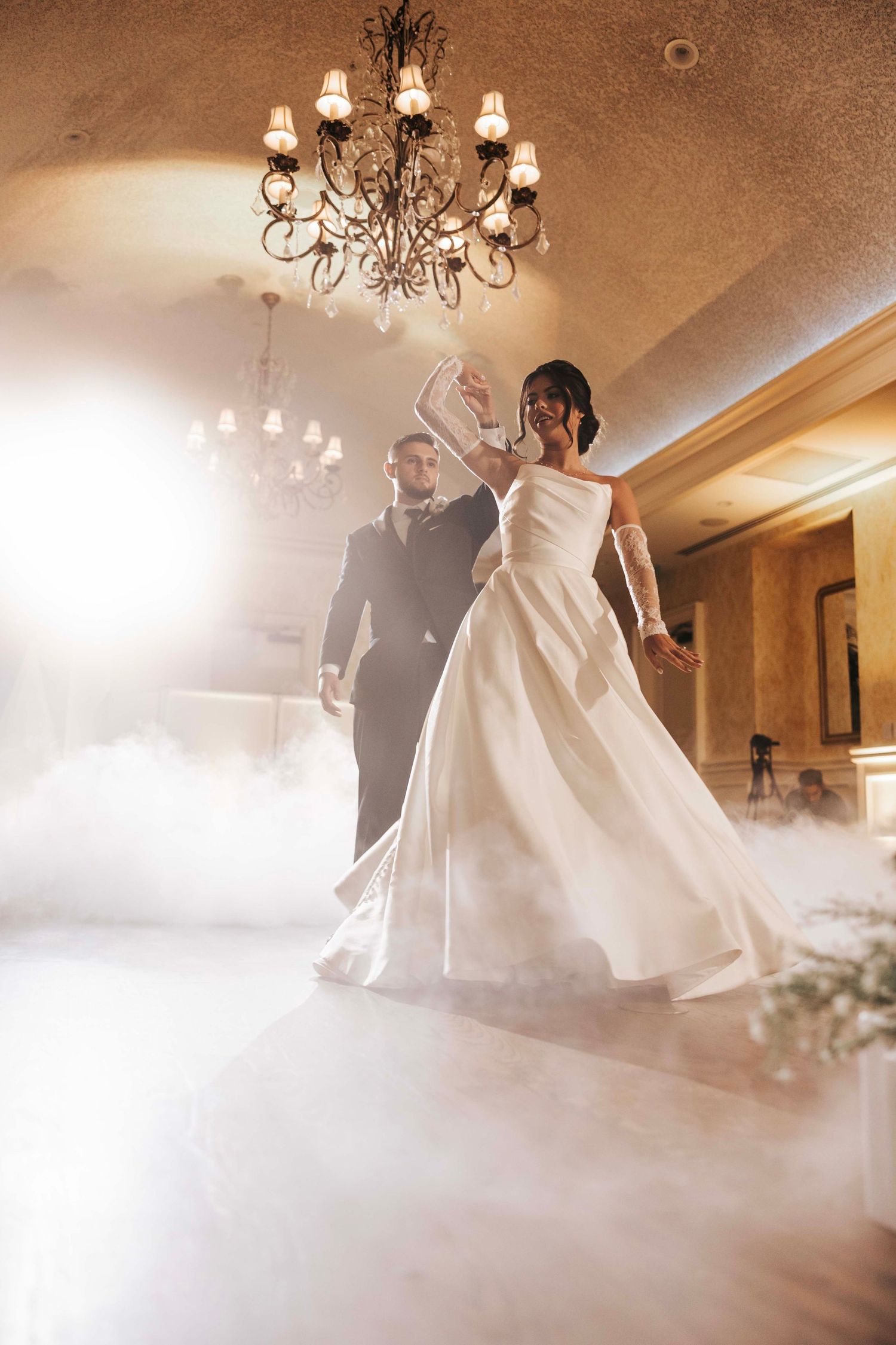 A couple shares their first dance under an elegant chandelier at their wedding reception with foggy atmosphere.