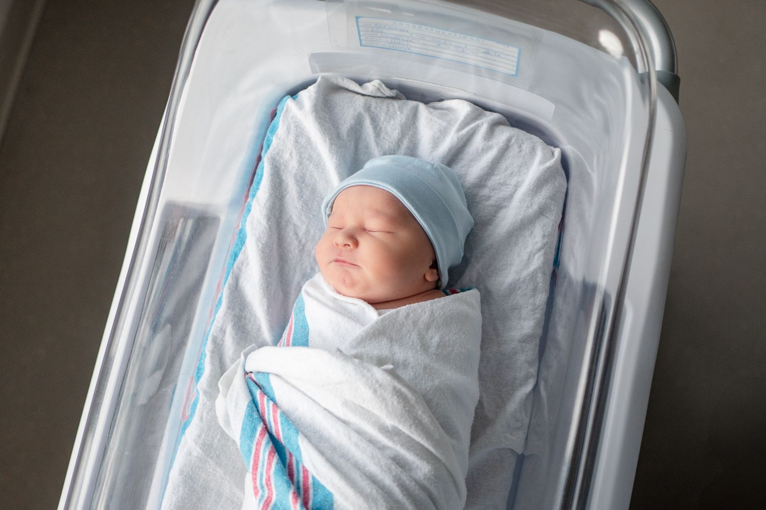 Newborn baby peacefully sleeping in hospital bassinet wrapped in white and blue striped blanket.