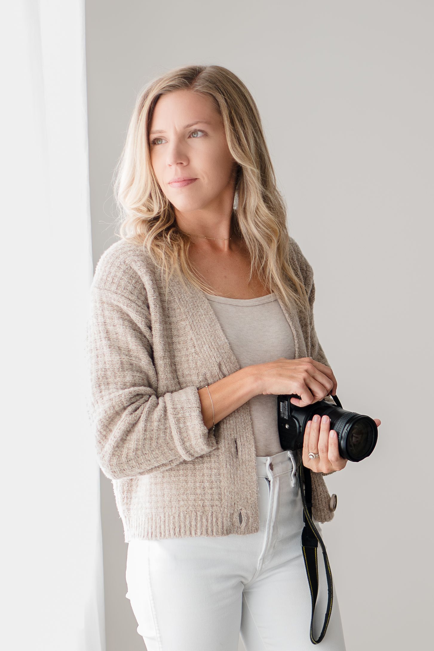 Professional photographer in beige cardigan and white jeans holding camera while standing by window in natural light.