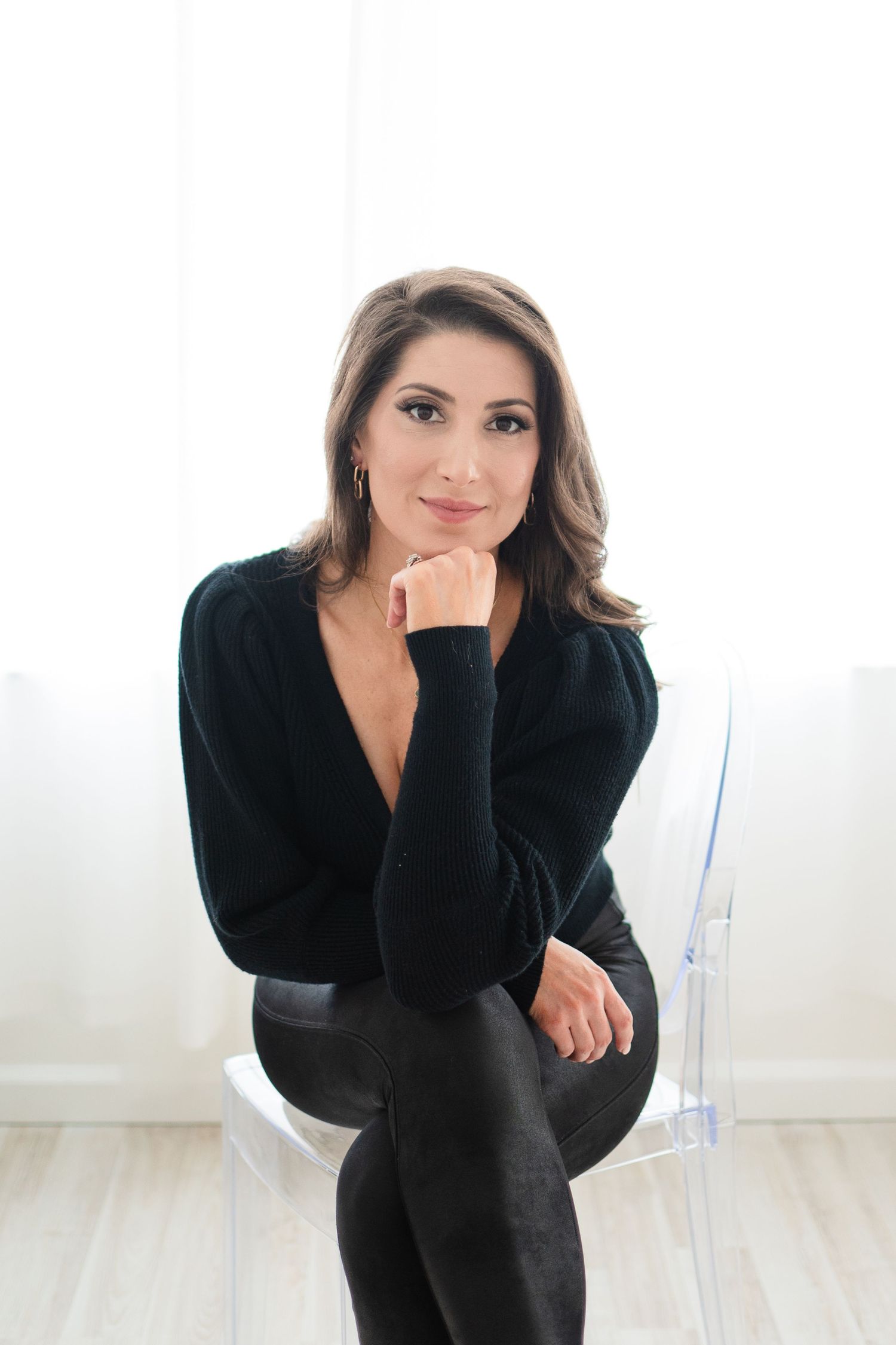 Professional portrait in a black outfit sitting on a clear chair against a white background.
