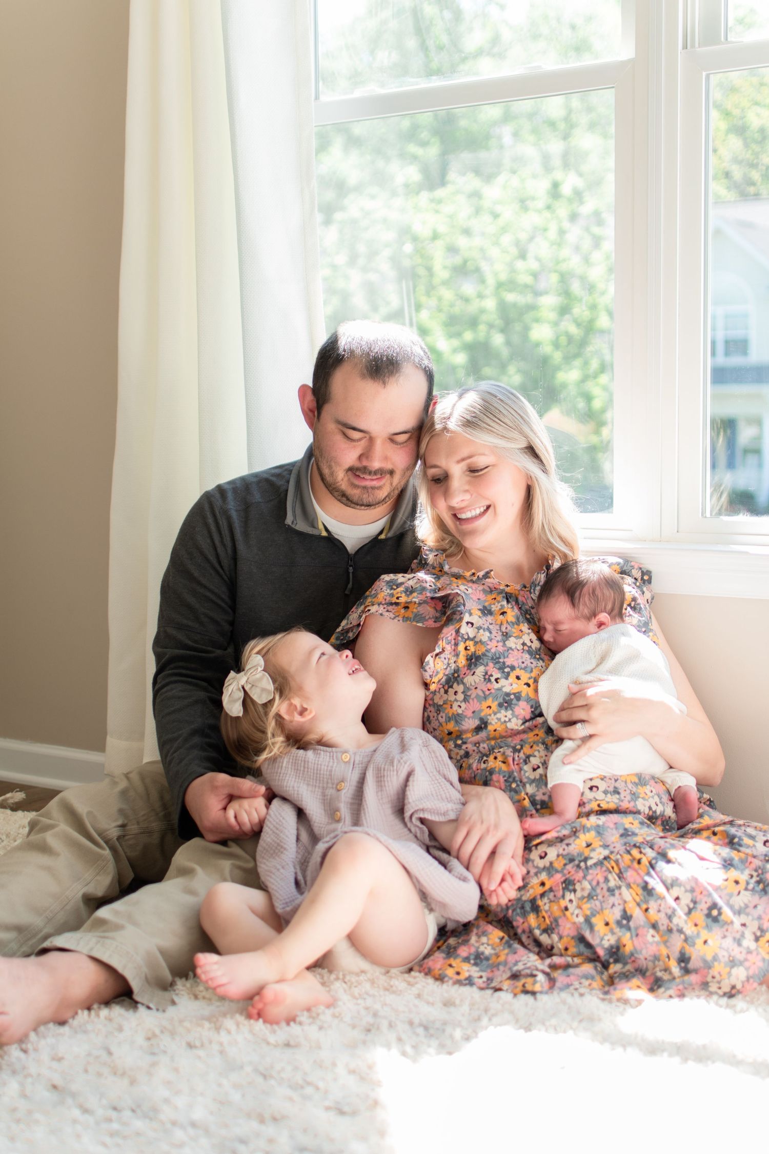 Intimate family portrait on plush carpet beside bright window with natural lighting.
