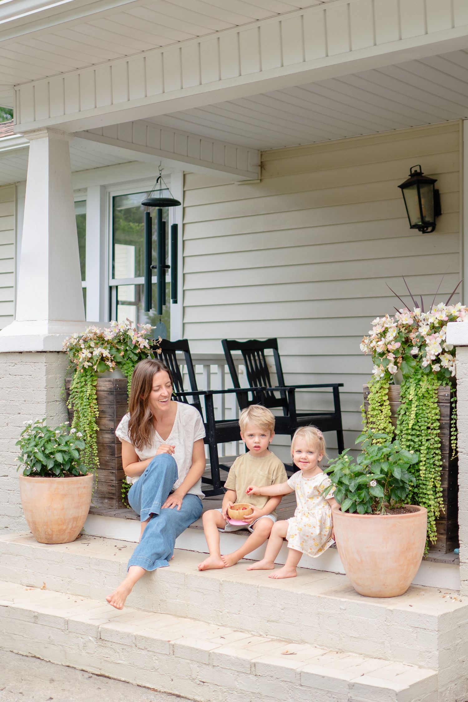 People enjoying a summer day relaxing on white farmhouse porch steps with rocking chairs and potted flowers.