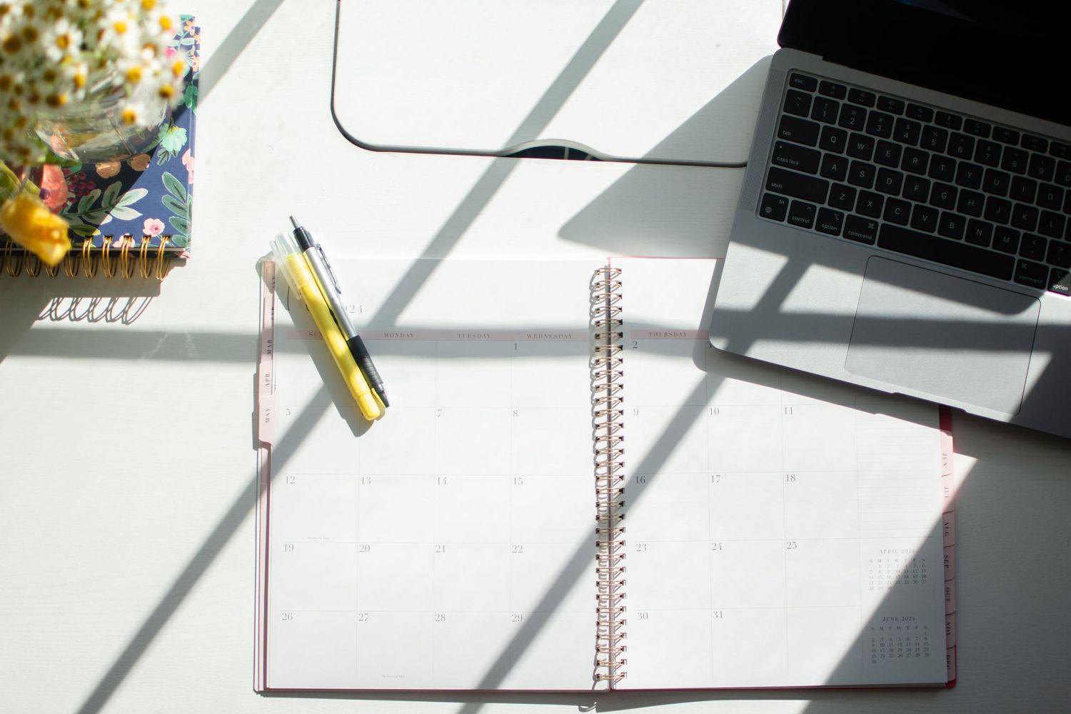 Open spiral notebook with laptop and stationery on white desk in sunlight.