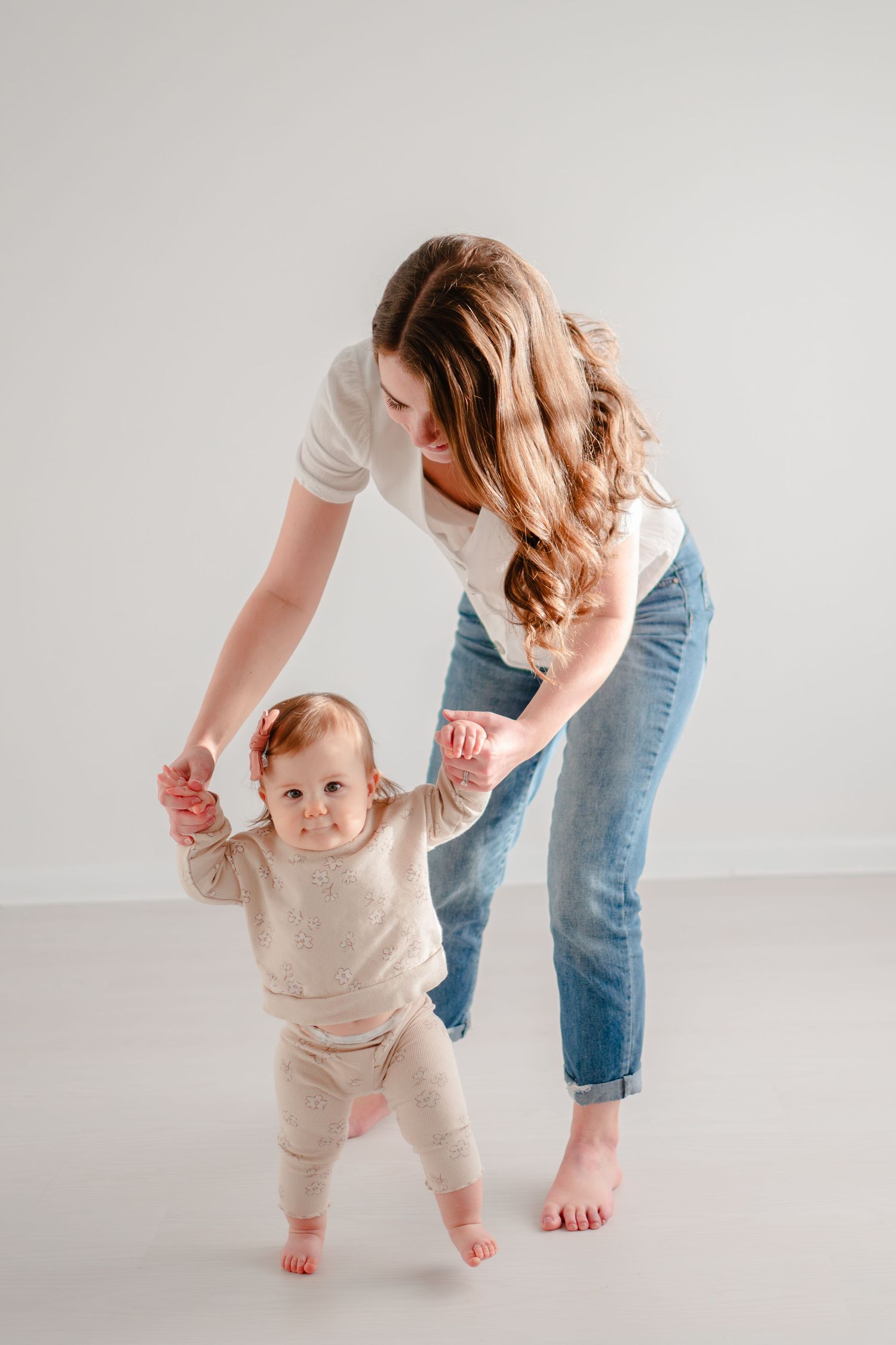 A parent in jeans helps their young baby take first steps on a white background while barefoot.
