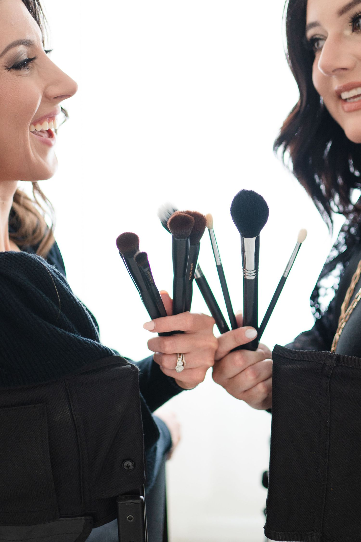 Two makeup artists sharing makeup brushes and tools while smiling and dressed in black.