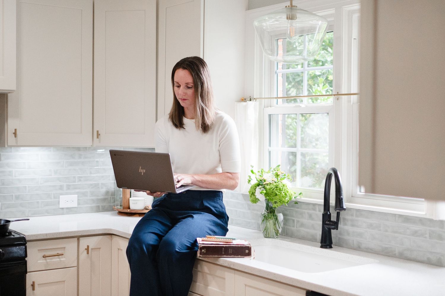 Person sits on white kitchen counter working remotely on laptop in bright modern kitchen with subway tile backsplash.