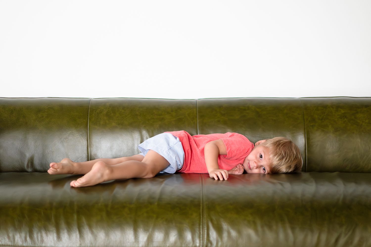 Child sleeping peacefully on dark green leather sofa in natural light.