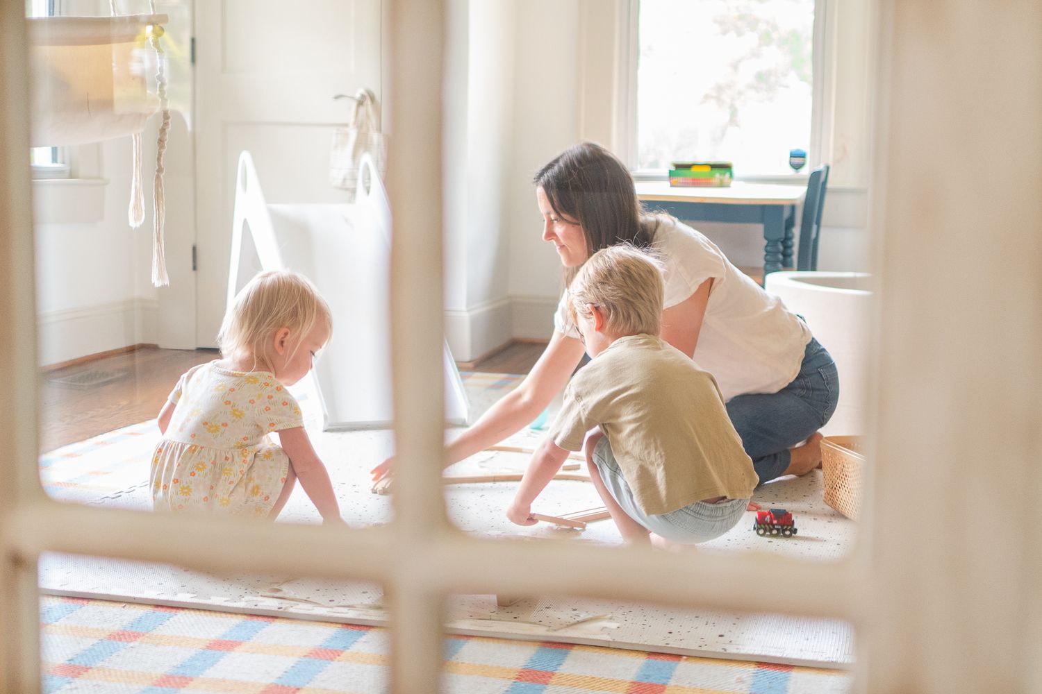 Through window bars, people clean floor together in sunlit room with colorful rug.