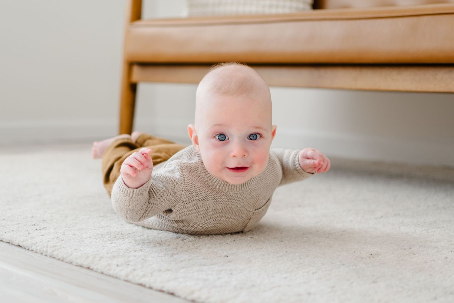 Baby wearing beige sweater lying on plush carpet near wooden furniture with arms outstretched and smiling.