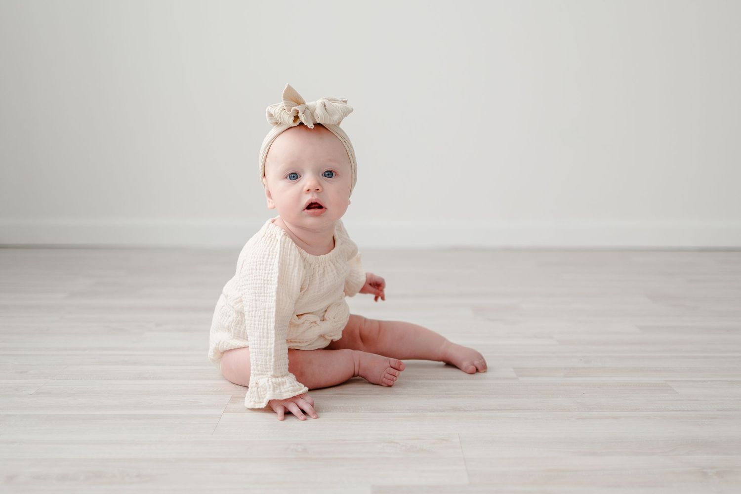Baby wearing white sweater and crown sits on light wooden floor in minimalist setting.