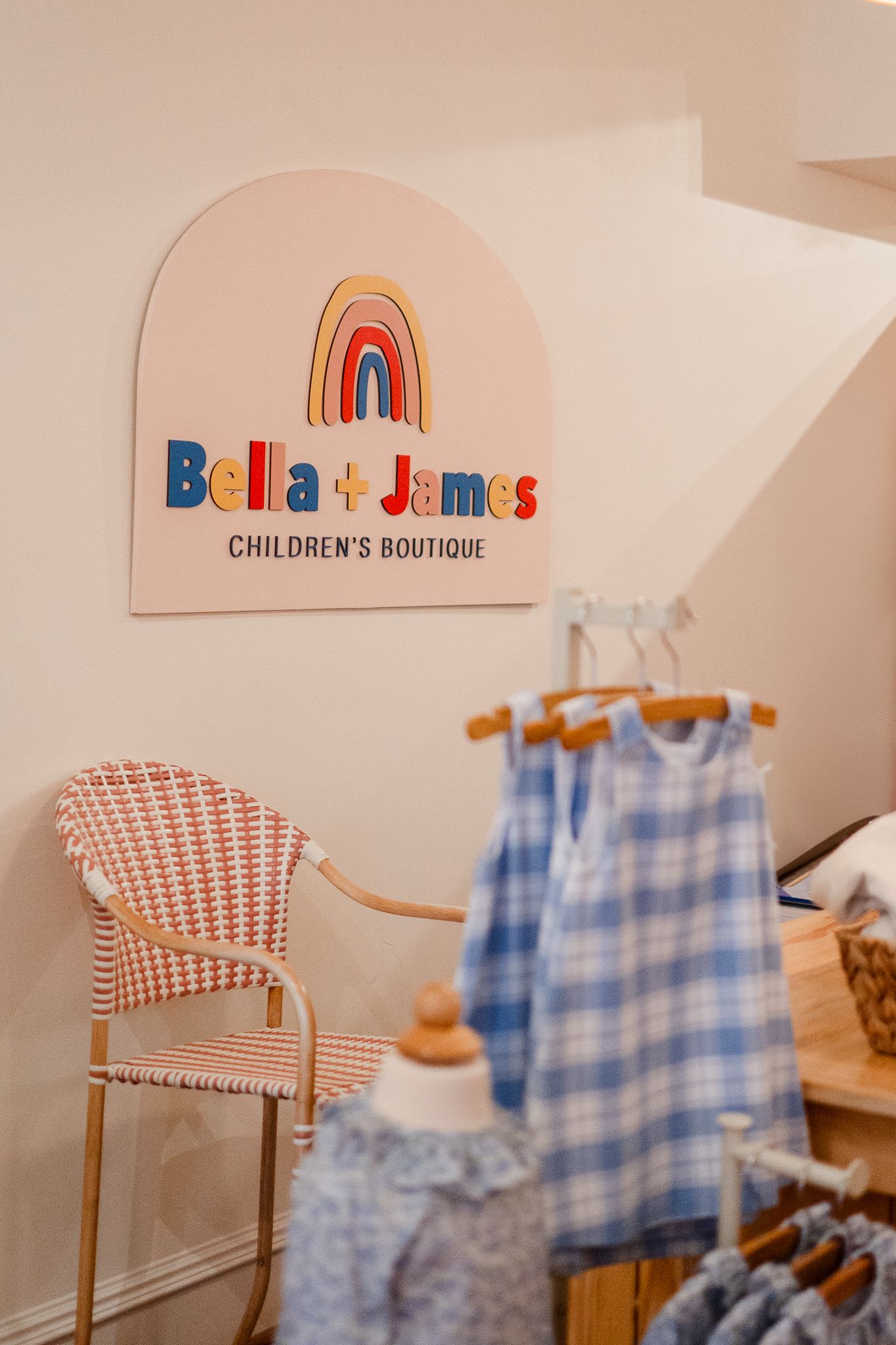 Rainbow logo of Bella + James children's boutique with pink chair and blue gingham dress displayed in store corner.