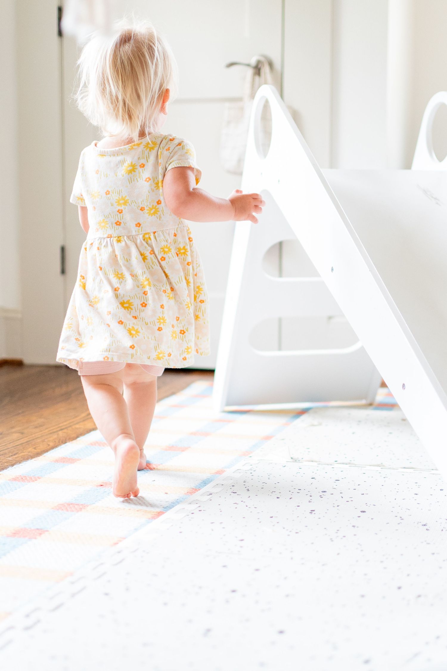 A toddler in a yellow floral dress walks barefoot on a white wooden floor near white shelving in a bright room.