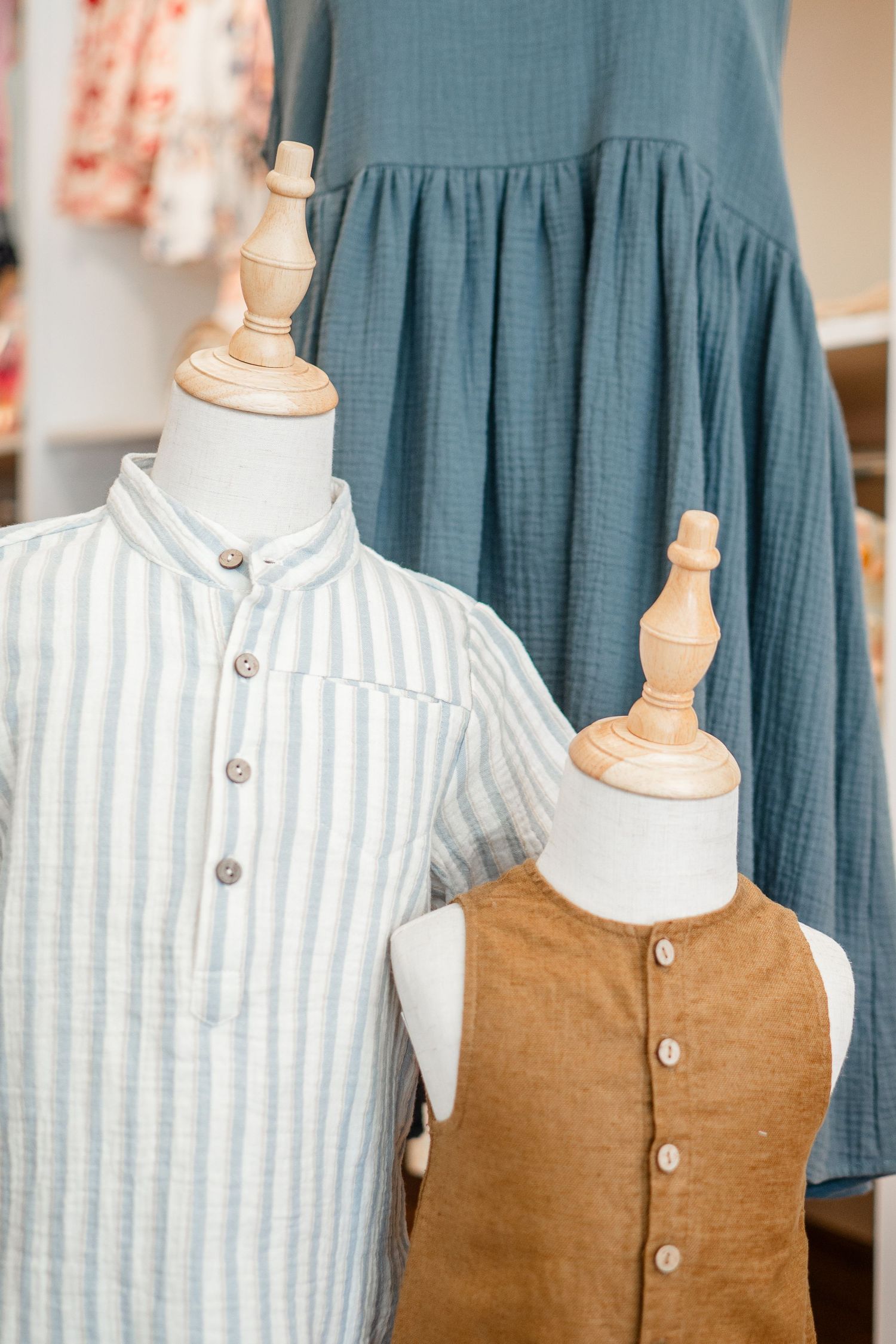 Three clothing mannequins display children's apparel including a striped shirt, brown vest and blue dress in natural tones.