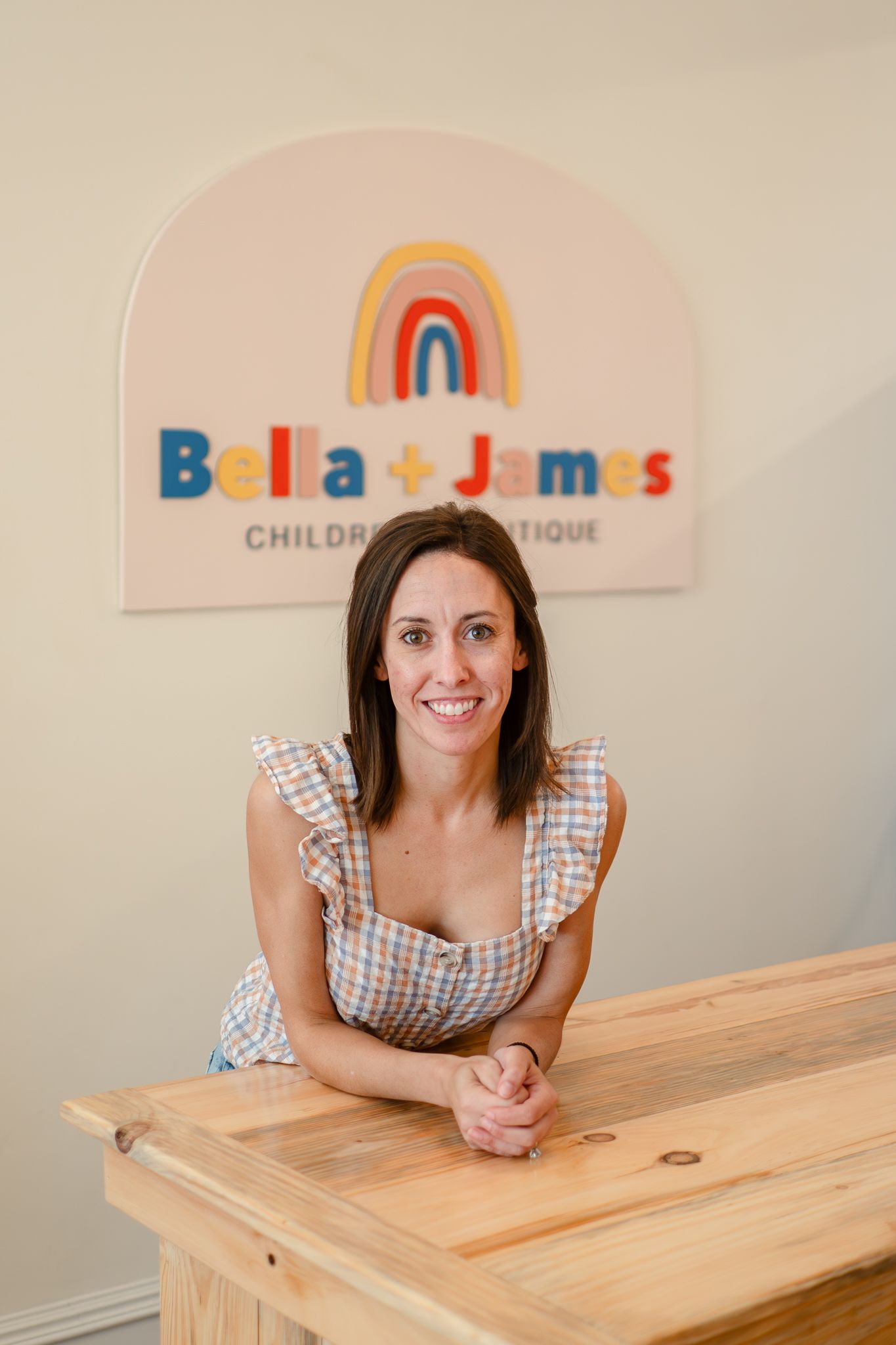 A woman in a gingham dress poses at a wooden table beneath a rainbow logo sign for a children's boutique.