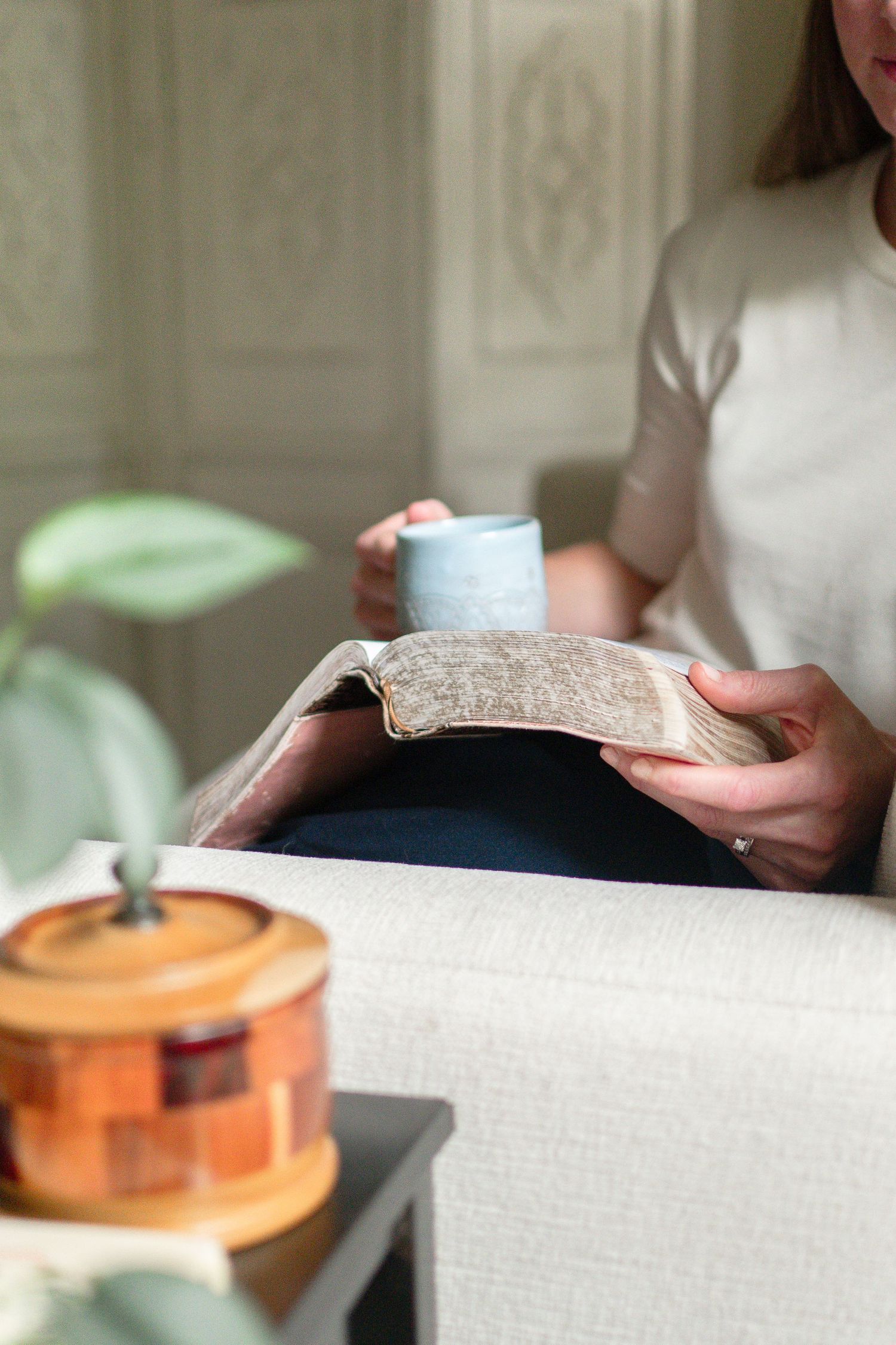 Someone relaxes on a couch with a book and blue mug while a ceramic jar sits nearby on a side table.