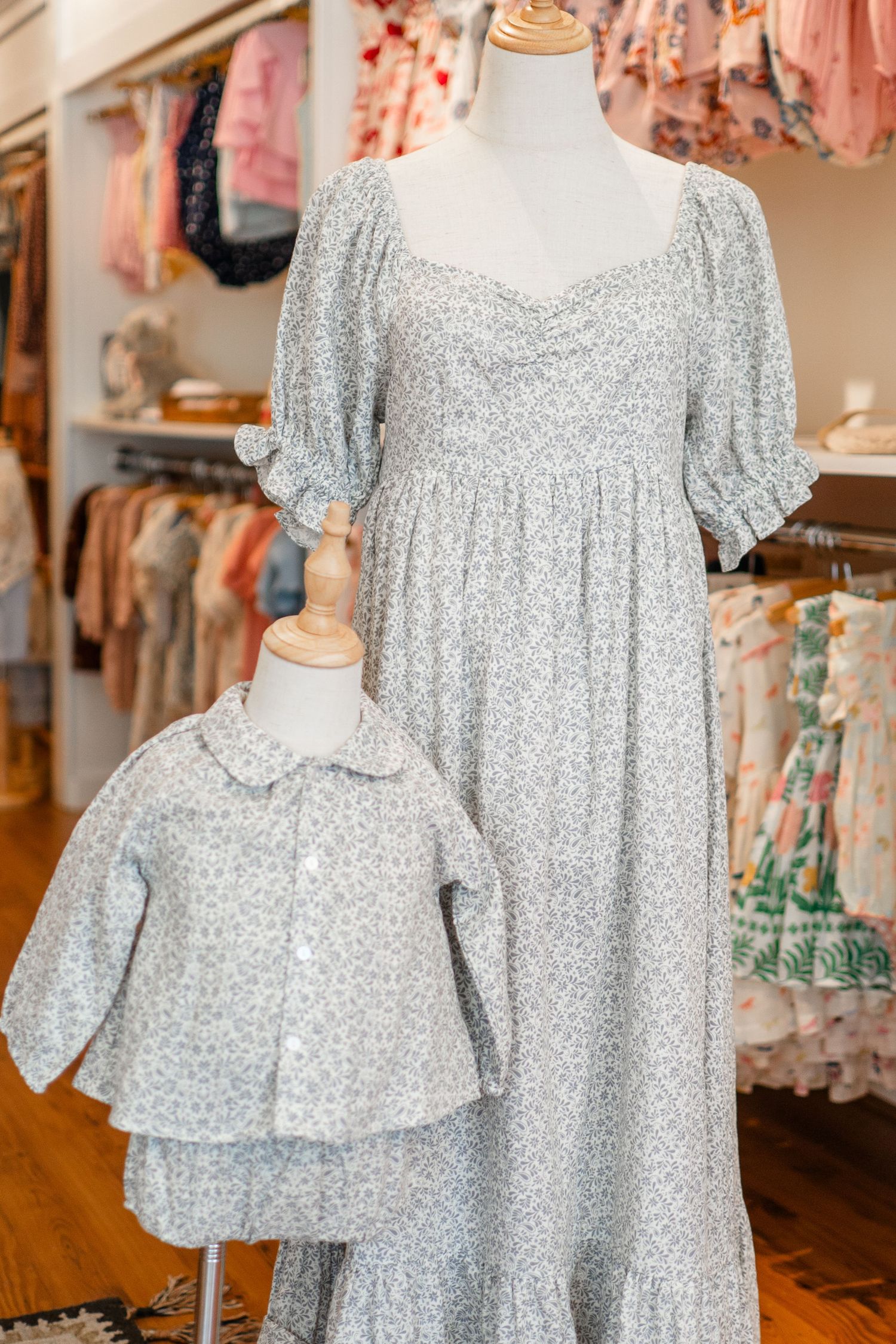 Matching floral print dress and toddler jacket in soft gray and white displayed on mannequins in a boutique clothing store.