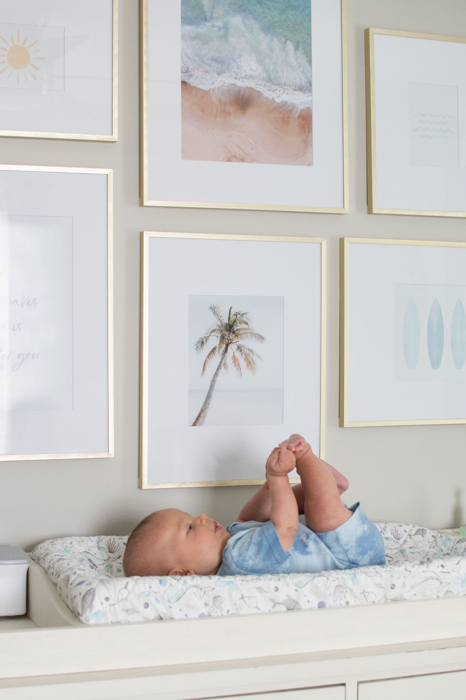 A baby lies on a changing table beneath a gallery wall featuring coastal and palm tree art prints in white frames.