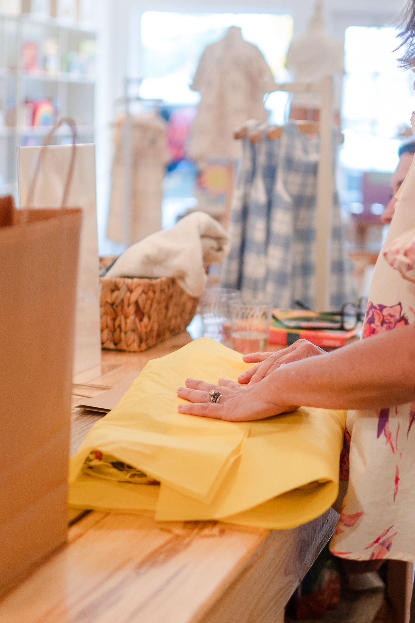 Hands fold yellow fabric on a wooden counter in a retail store with shopping bags nearby.