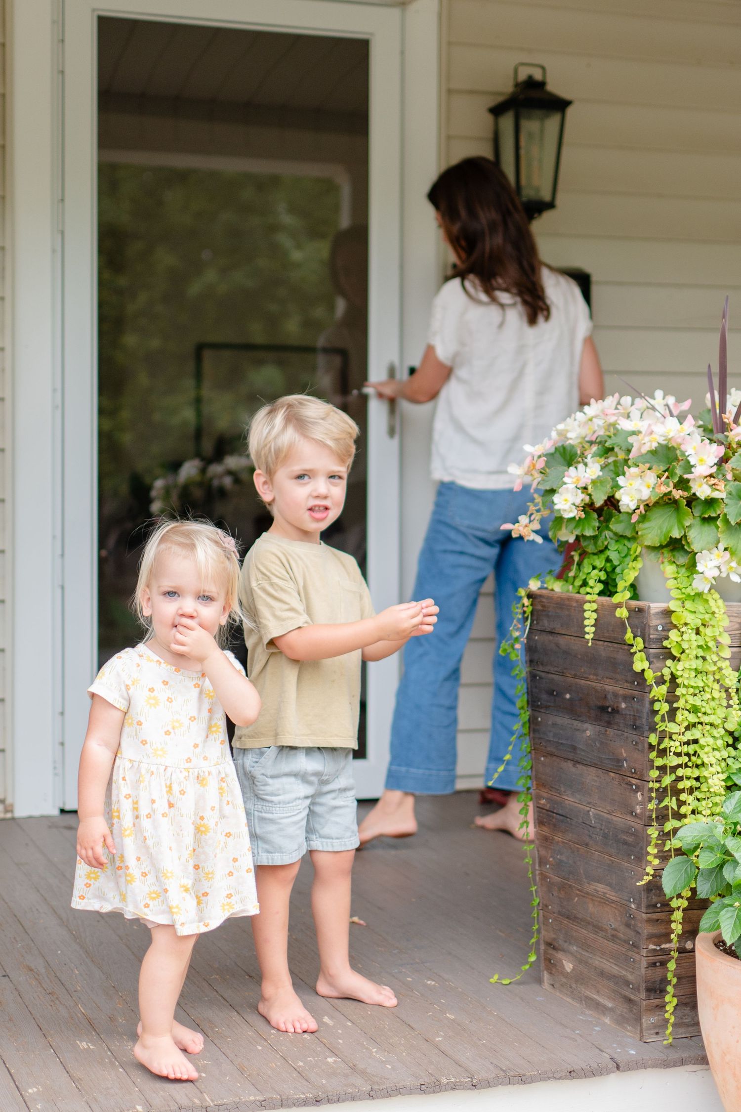 Children stand barefoot on a farmhouse porch while someone opens the front door, with a floral planter nearby.