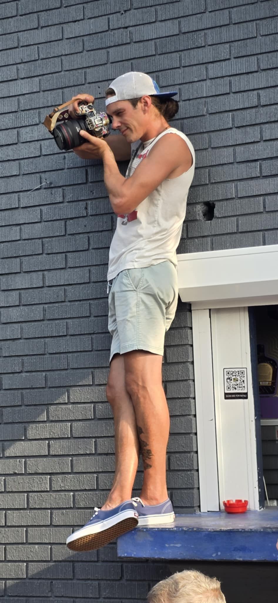 Person in casual summer attire with white baseball cap stands on ledge taking photos with camera against gray brick wall.