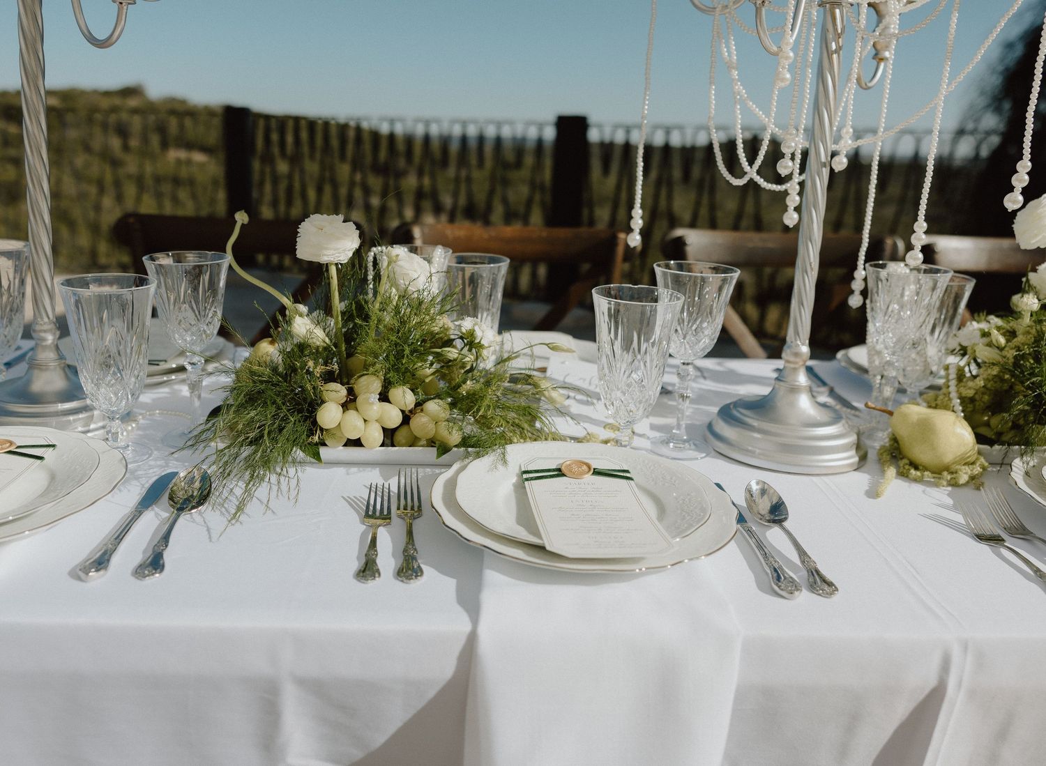 Elegant wedding table setting with white linens, crystal glassware and fresh greenery centerpiece at an outdoor venue.