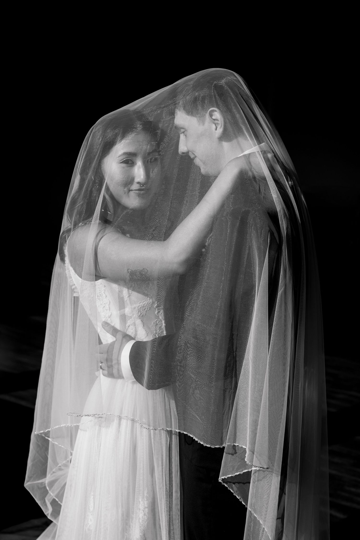 A romantic black and white portrait of a couple embracing under a delicate bridal veil.