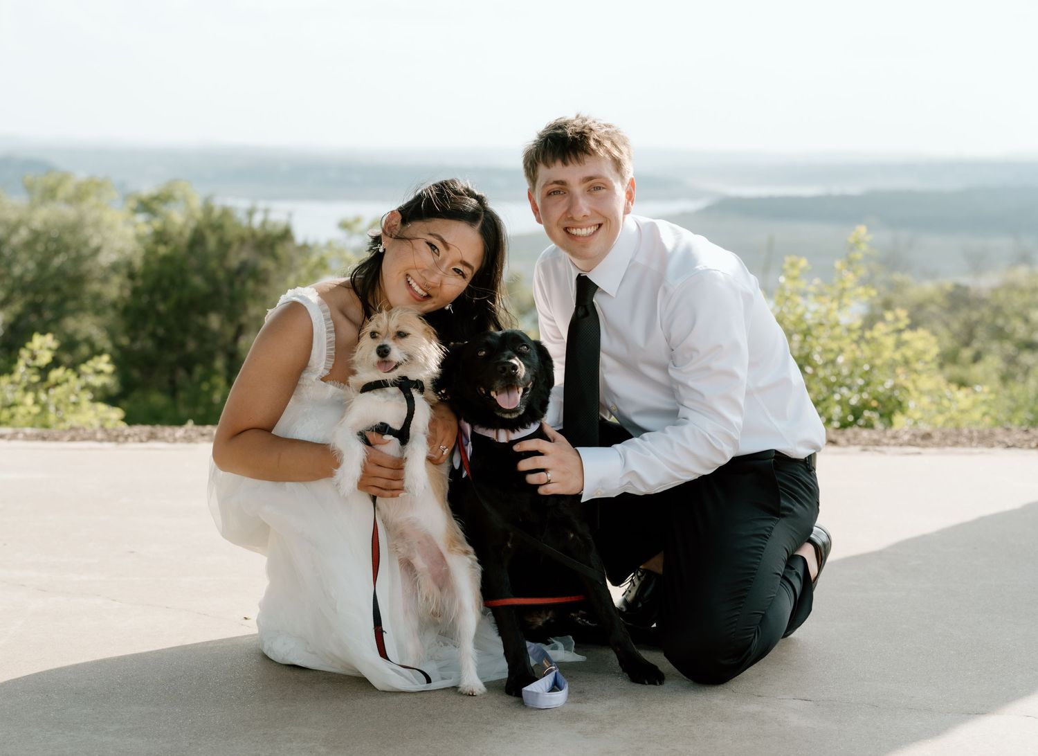 Young couple poses outdoors on a scenic overlook with their two small dogs on a sunny day.