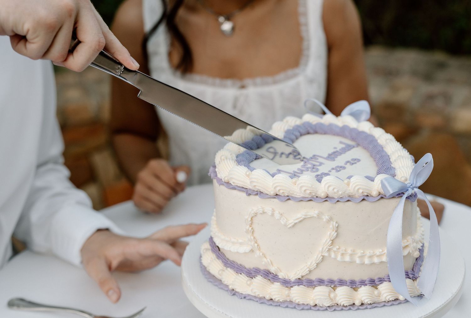 A decorative white cake with lavender trim and bow being cut with a silver knife at a celebration.
