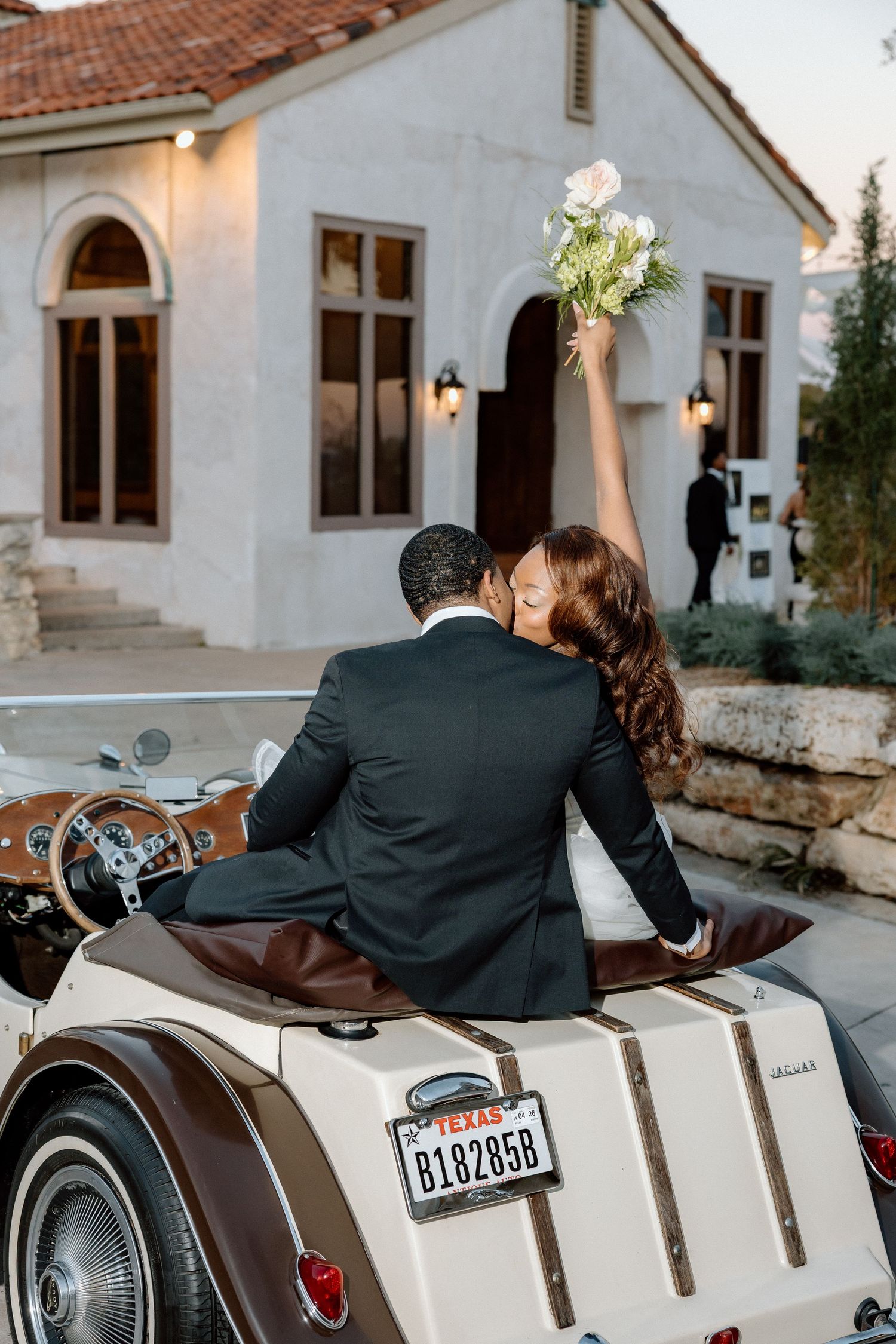 A couple sits on a vintage white motorcycle outside a Mediterranean-style building with arched doorways and sconces.