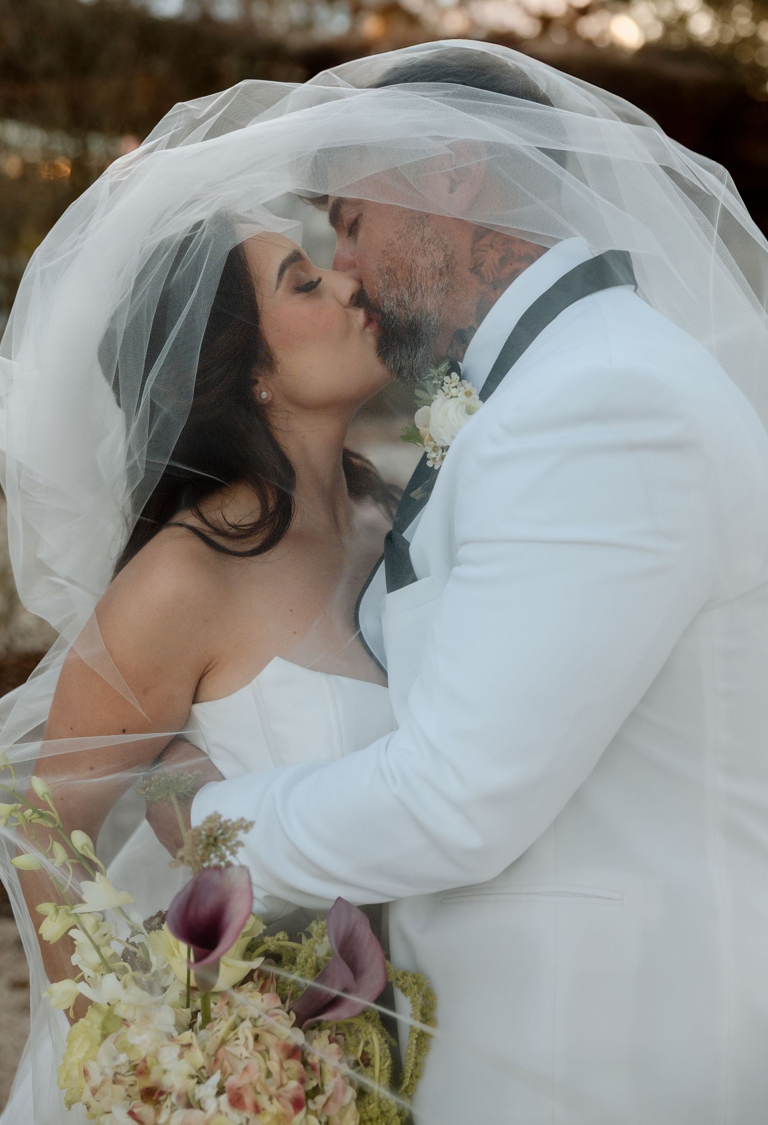 A romantic wedding kiss between the bride in white and groom in light colored suit beneath a flowing veil at sunset.
