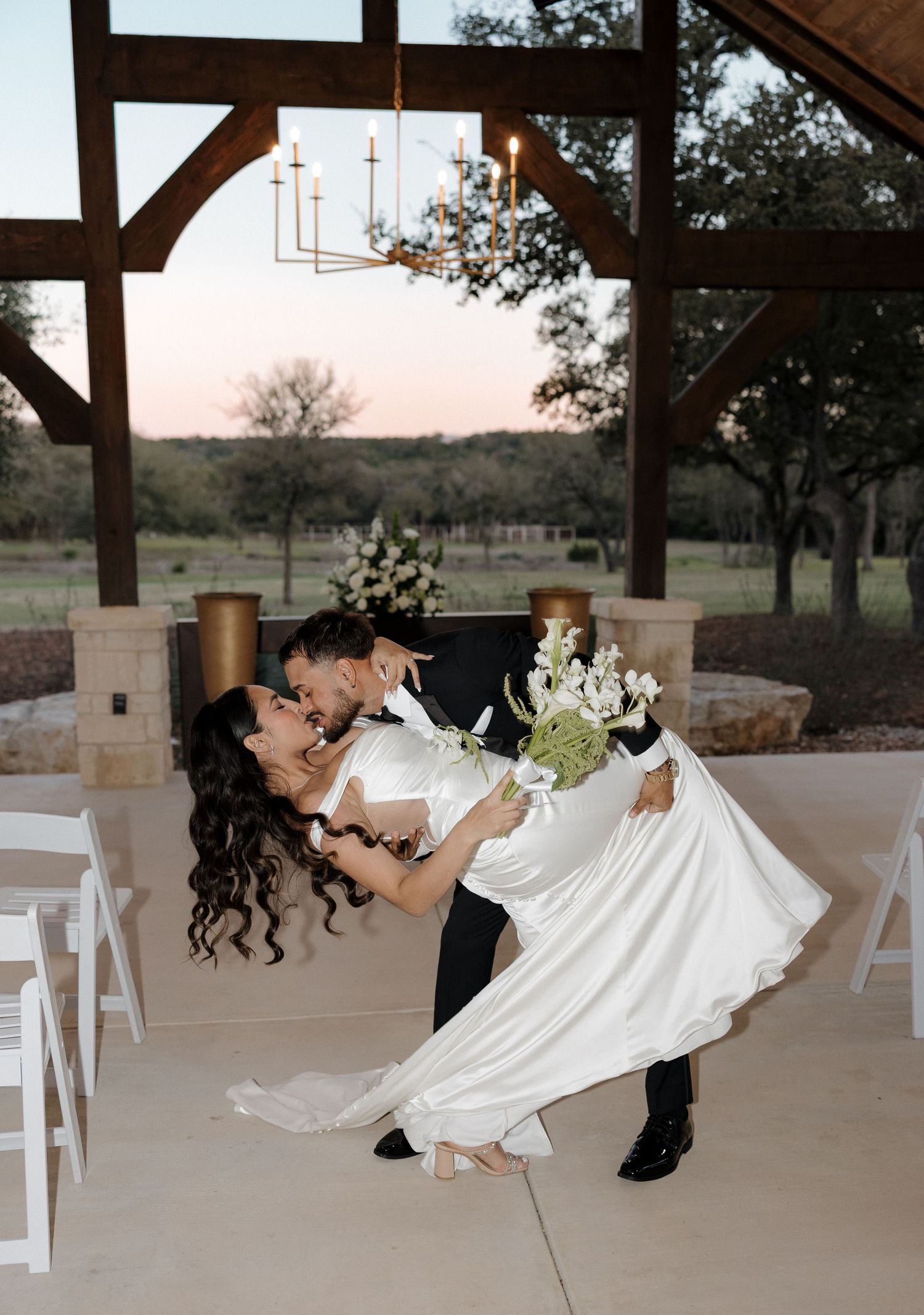 A romantic dip dance pose at an outdoor covered venue with a sunset view through wooden beams and white chairs.