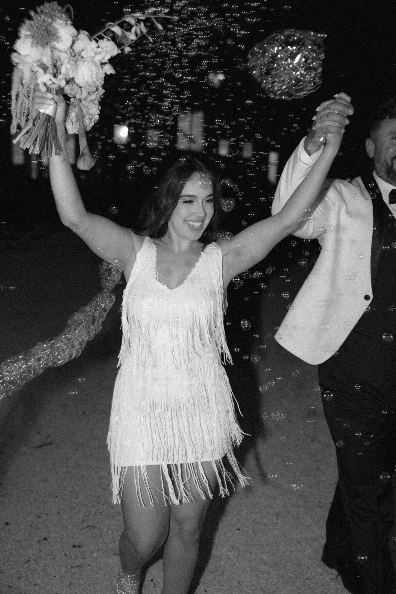 A celebratory moment showing someone dancing in a white fringed flapper-style dress while raising their arms at a party.