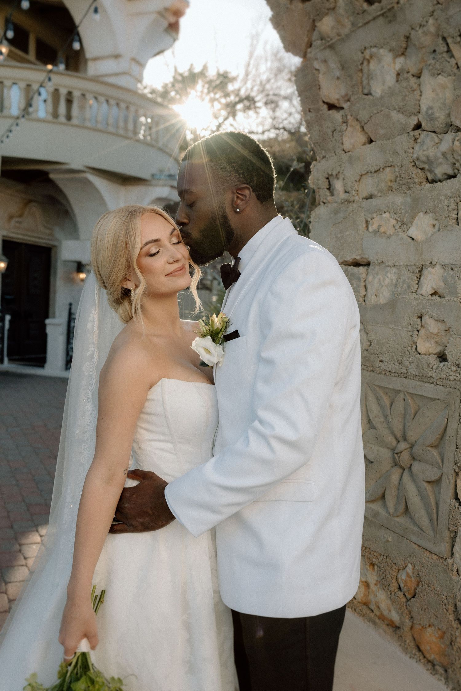 A romantic wedding couple embraces against a stone wall at sunset, the bride in white and groom in a white jacket.